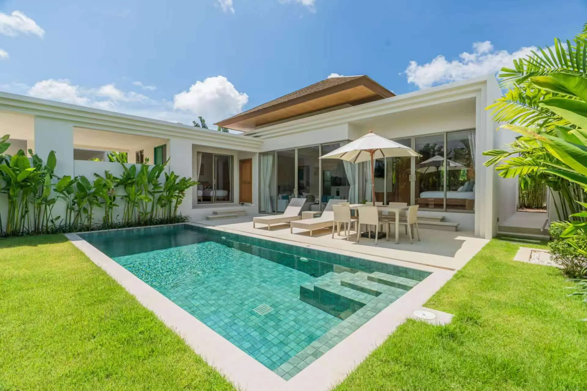 Private pool set in a lawn, with sun loungers and a shaded dining table on the terrace of a single-storey villa