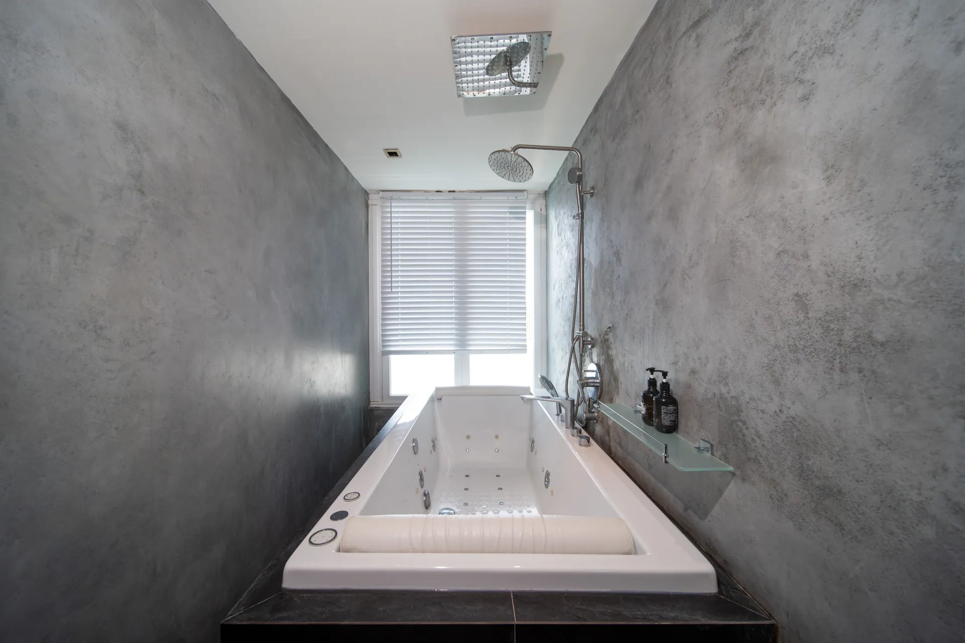 Bathroom with a deep bathtub and a rain shower between concrete-finish walls
