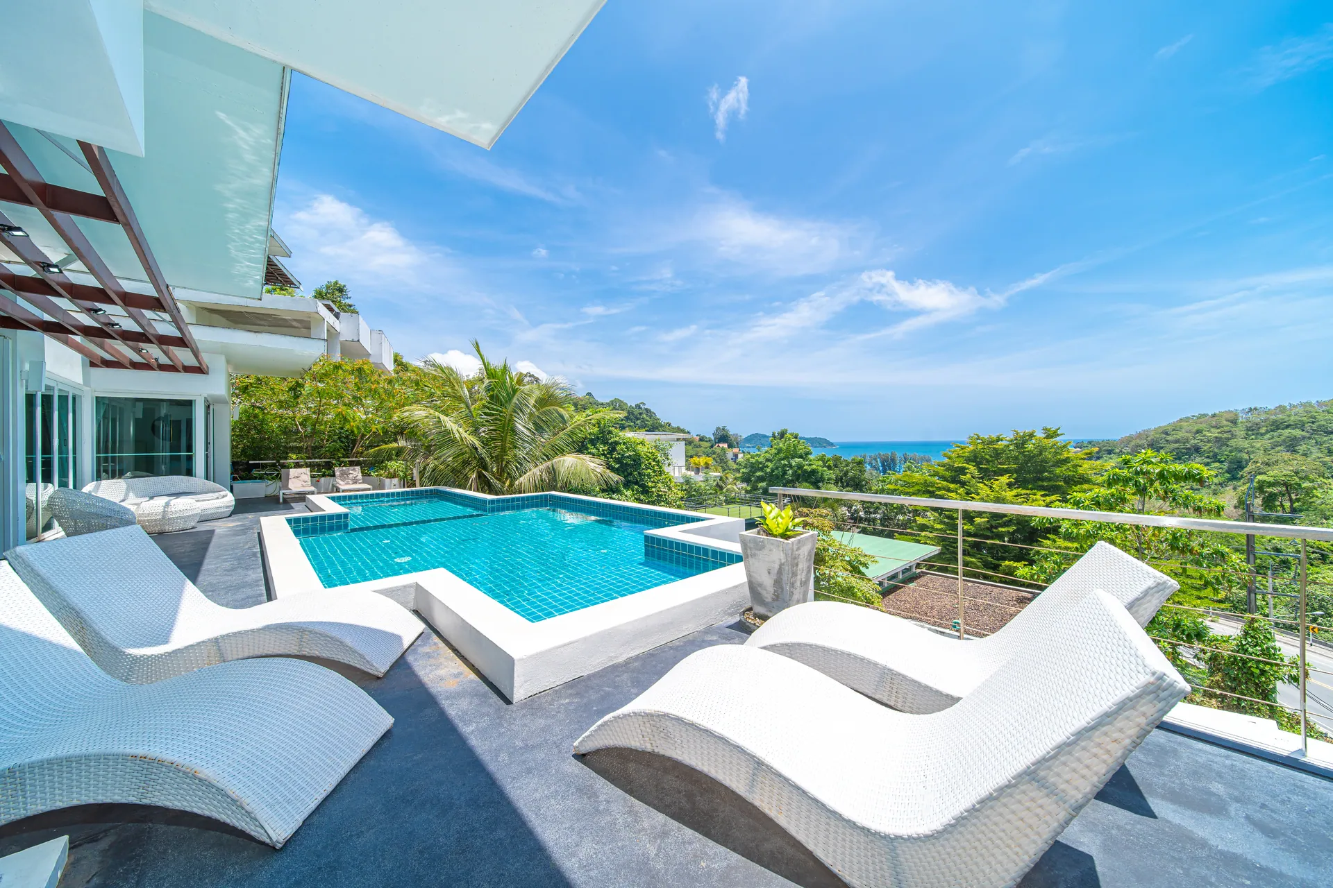 White sun loungers beside the pool, with trees and the sea beyond the terrace railing
