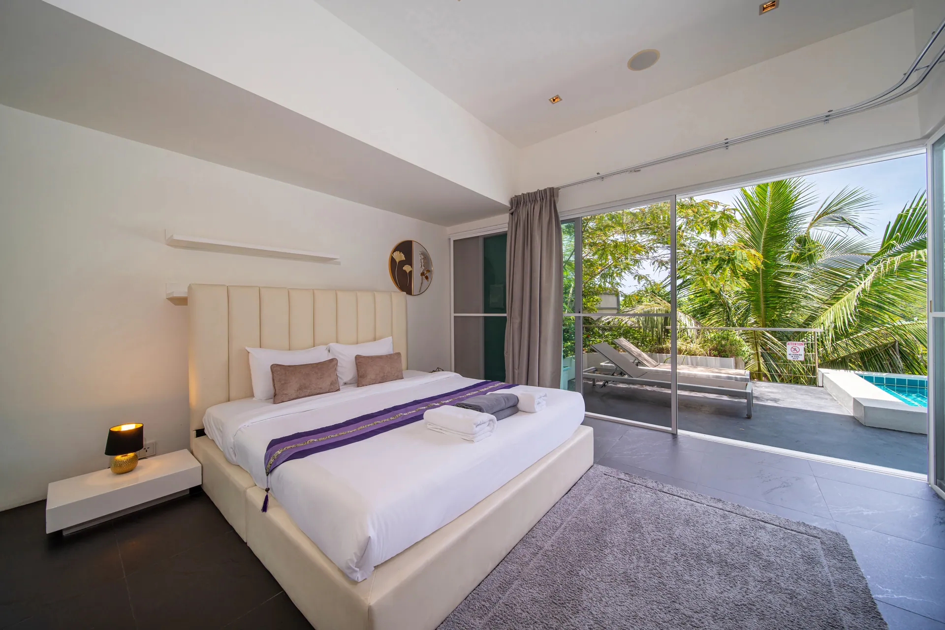 Bedroom with a cream headboard and glass doors to a balcony, with palms and the pool beyond