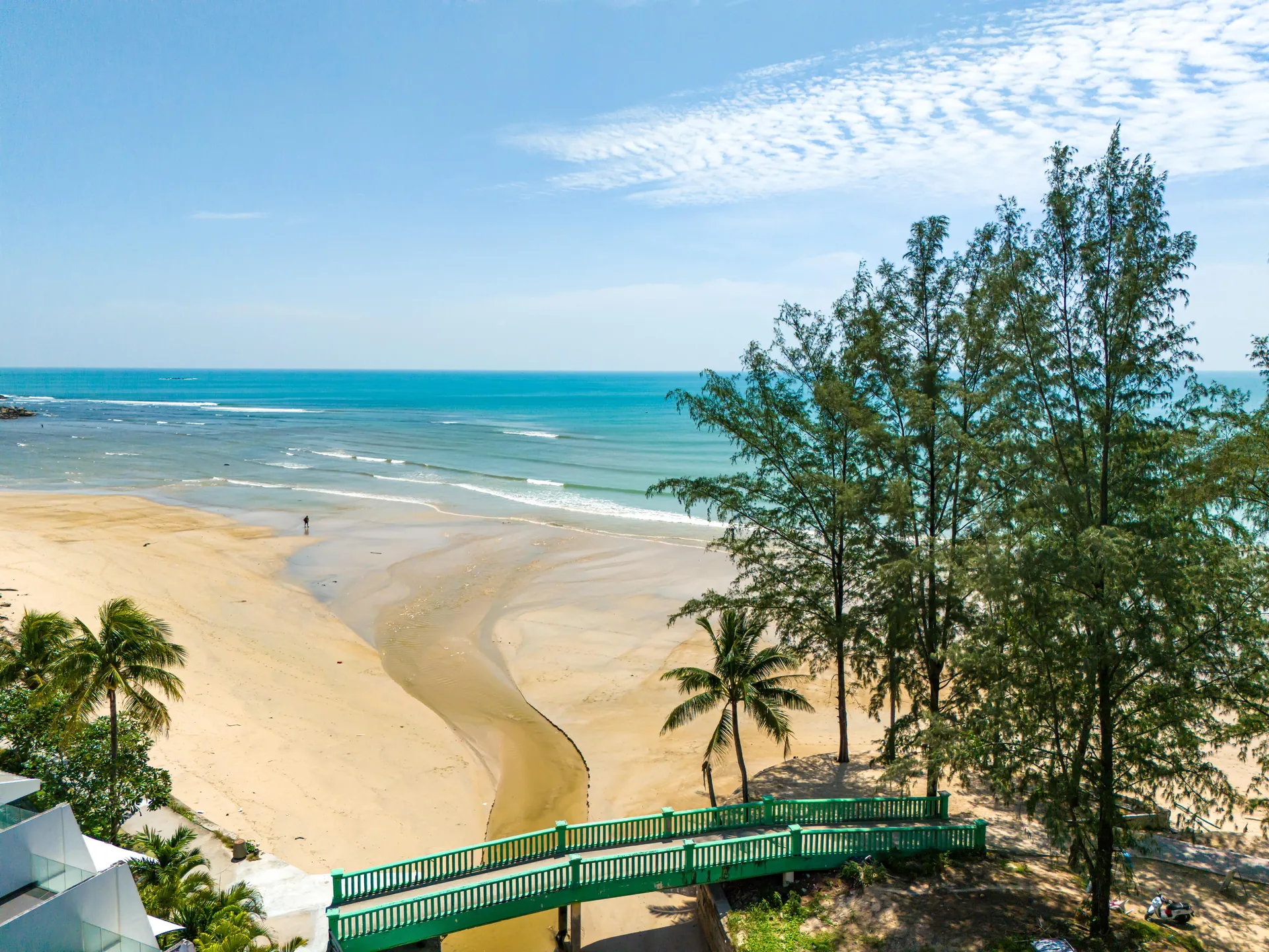 Kamala Beach at low tide, framed by casuarina trees and a green footbridge
