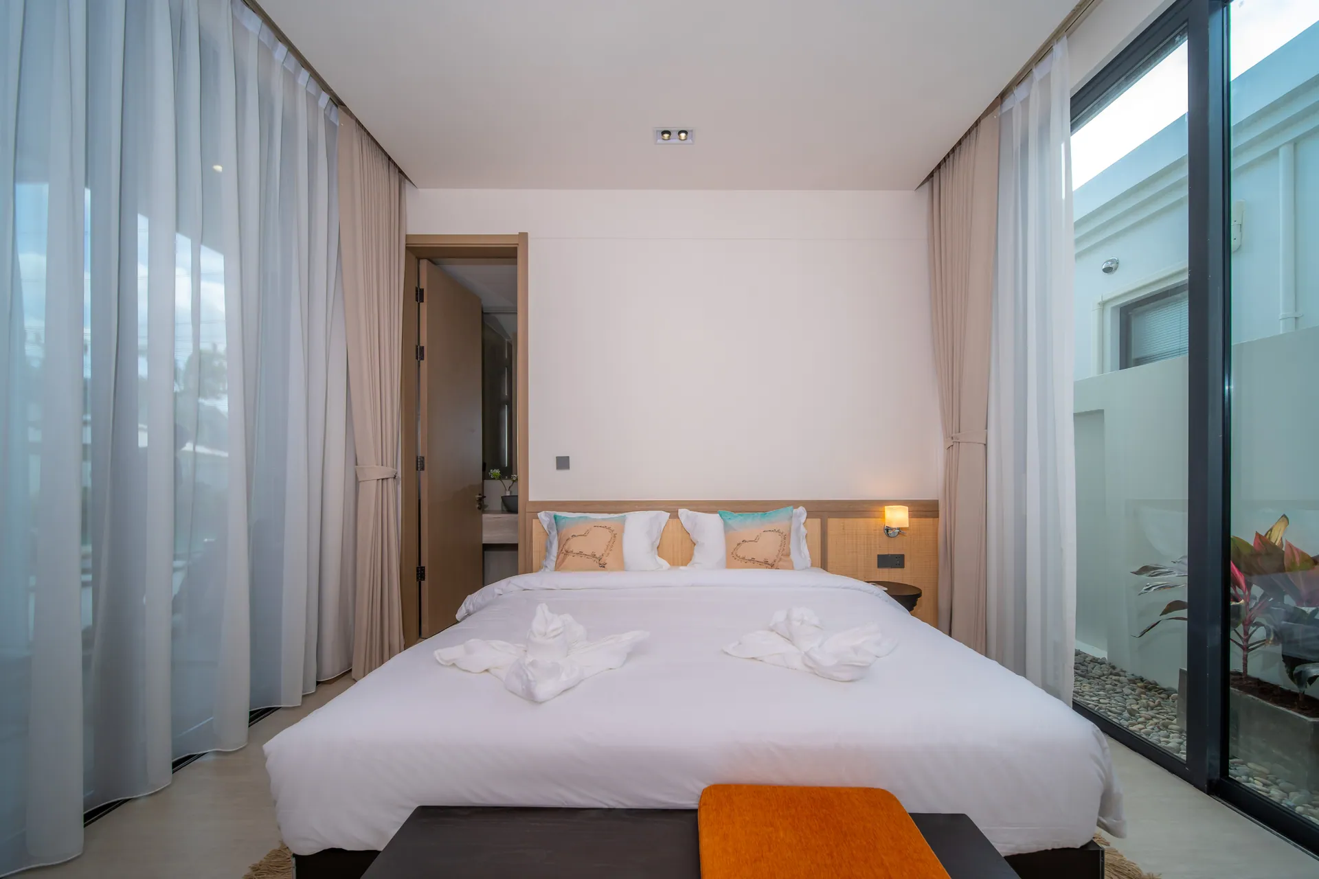 Bedroom with a king bed between sheer curtains and a glass wall onto a small planted courtyard