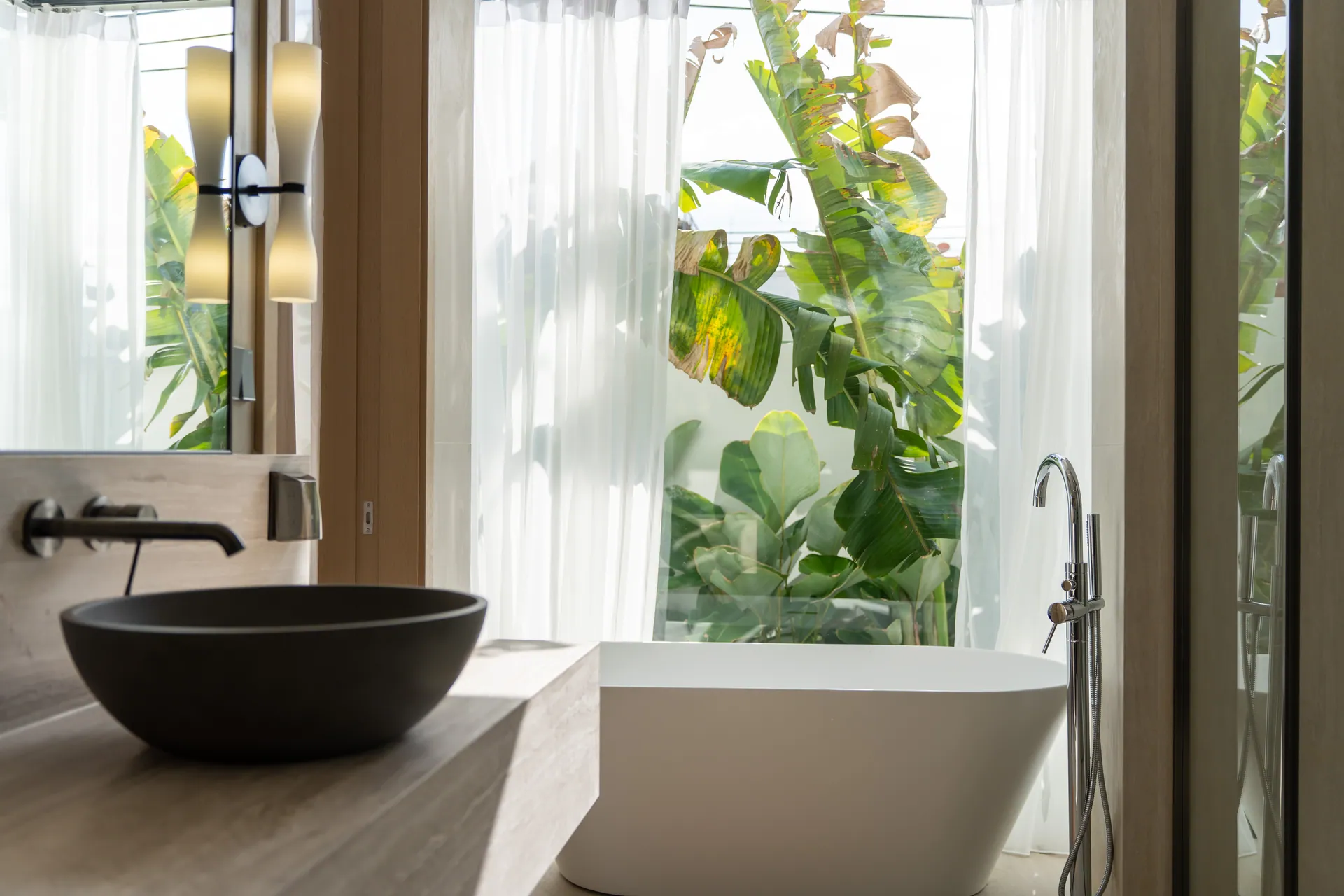Freestanding bath and a stone basin beside a window framed by tropical plants