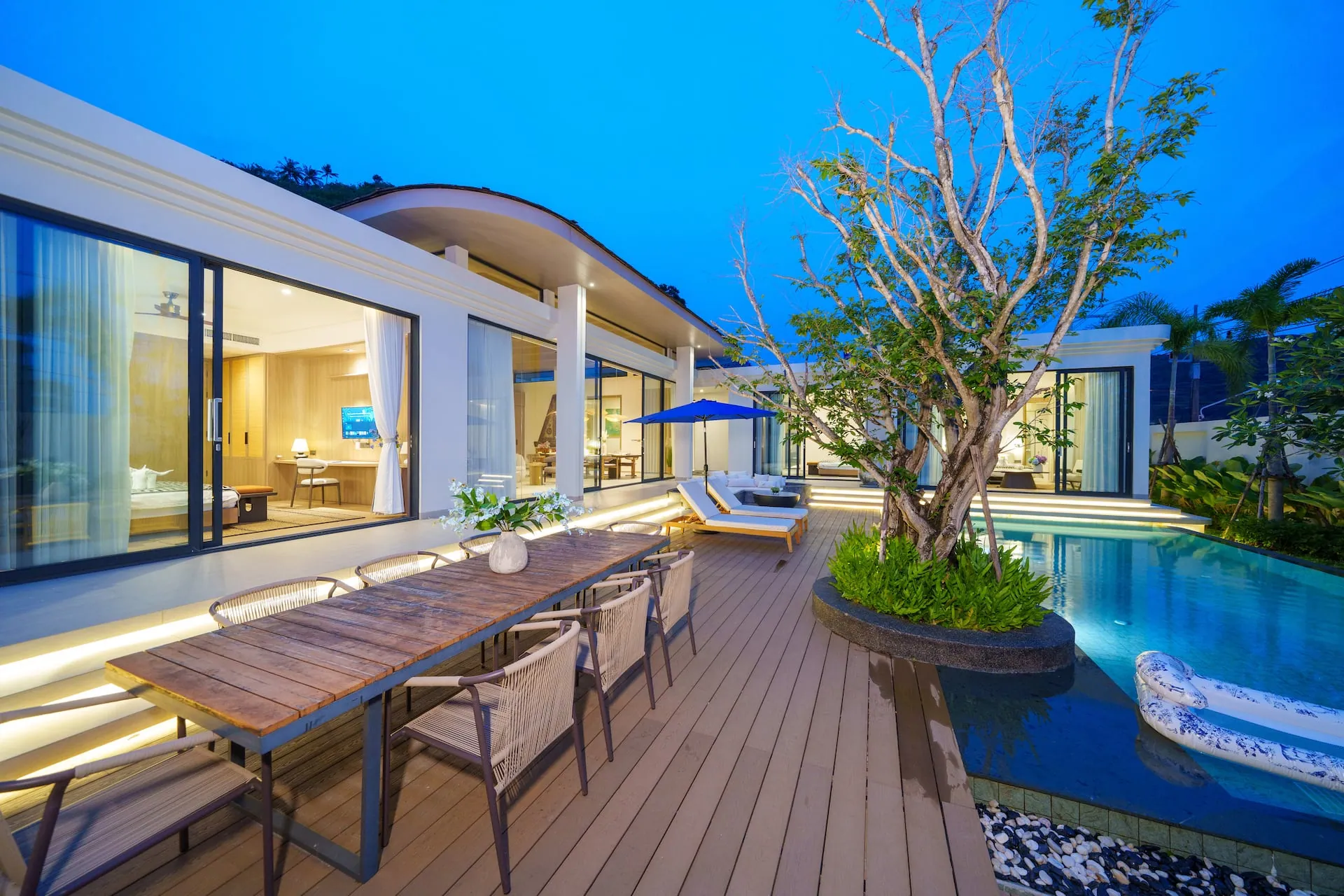 Outdoor dining table on the deck beside the pool and a courtyard tree at dusk