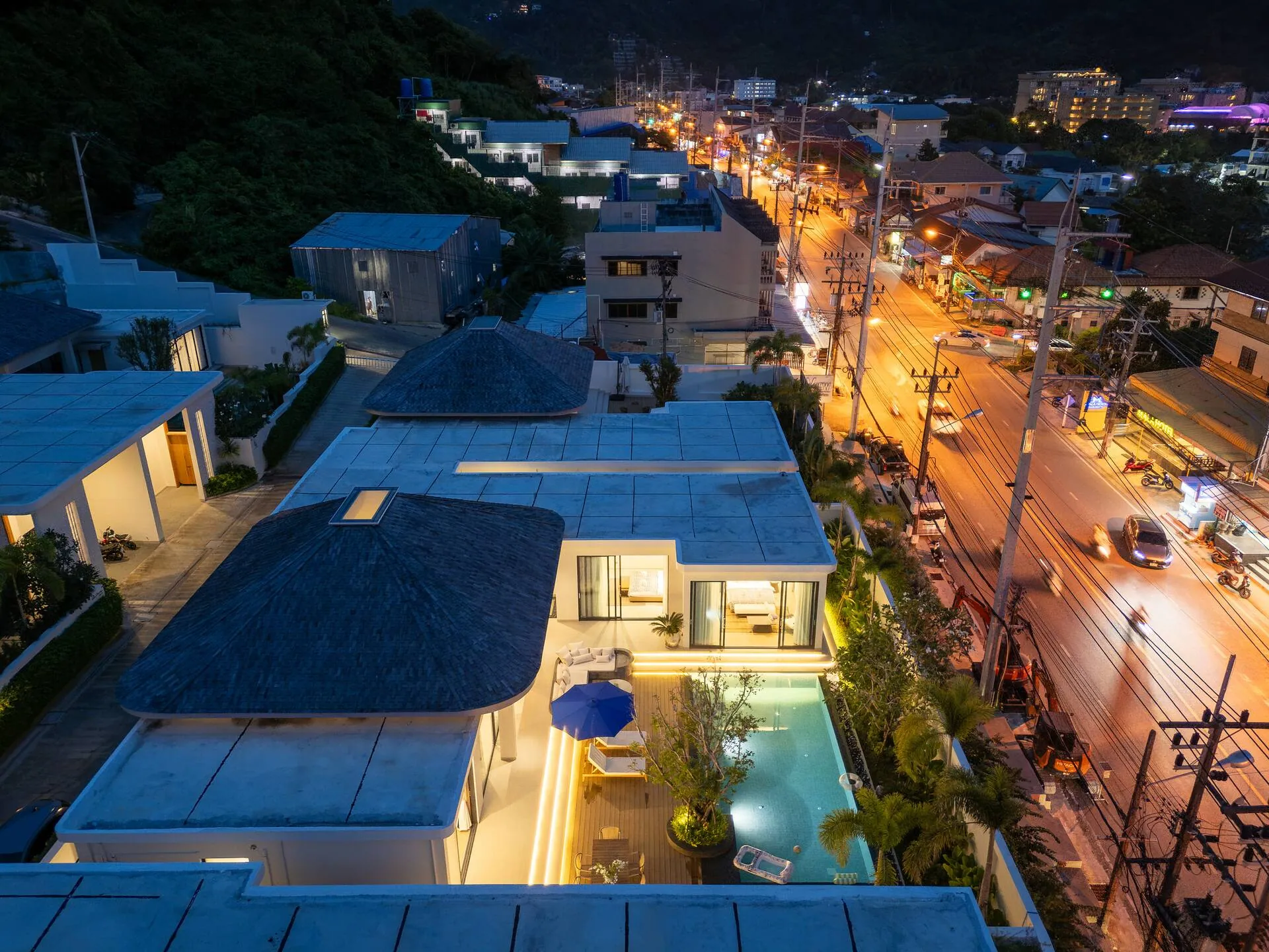Aerial view at dusk over the villa, its lit pool and the surrounding streets and hillside