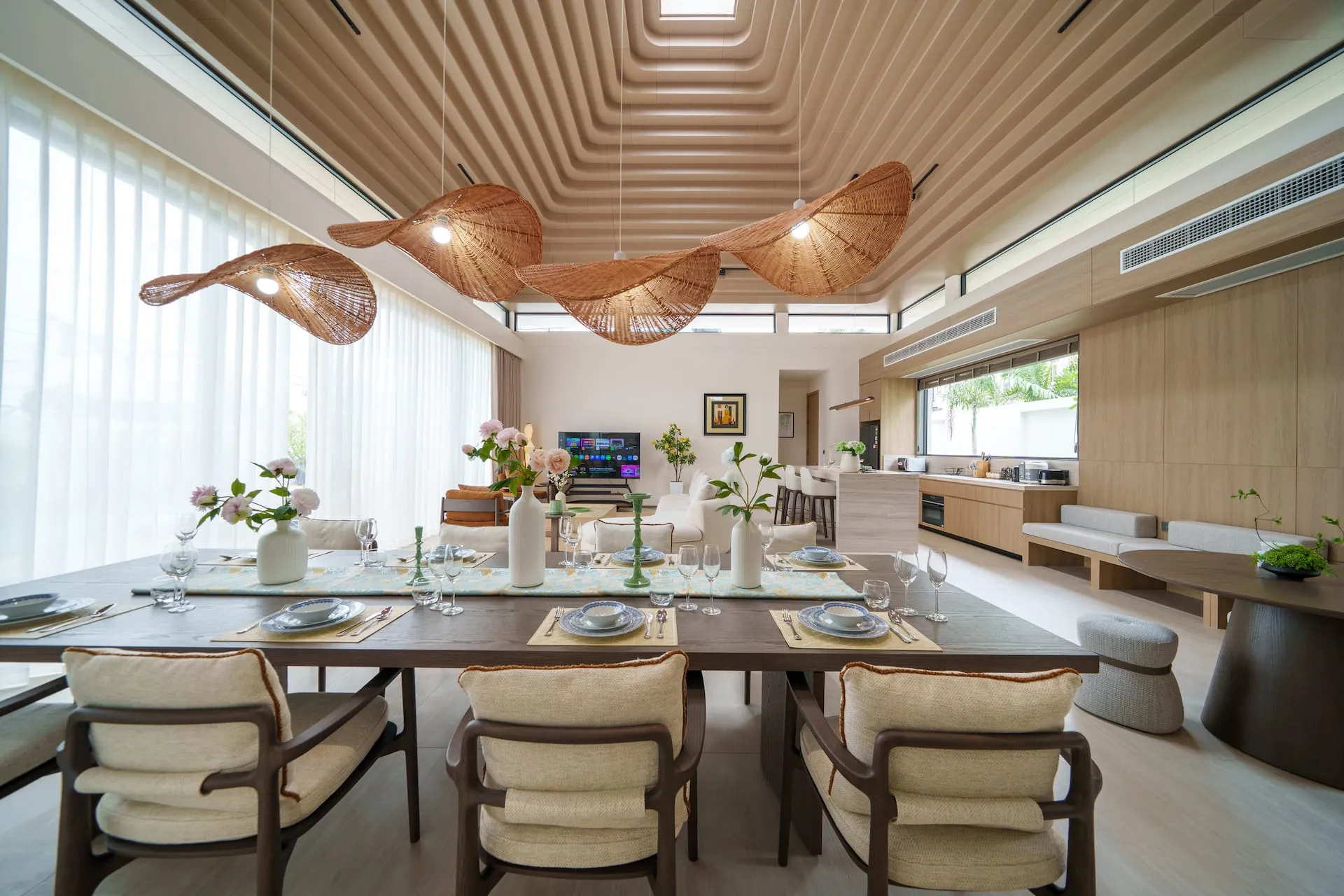 Long dining table laid beneath woven pendant lights, with the living area beyond
