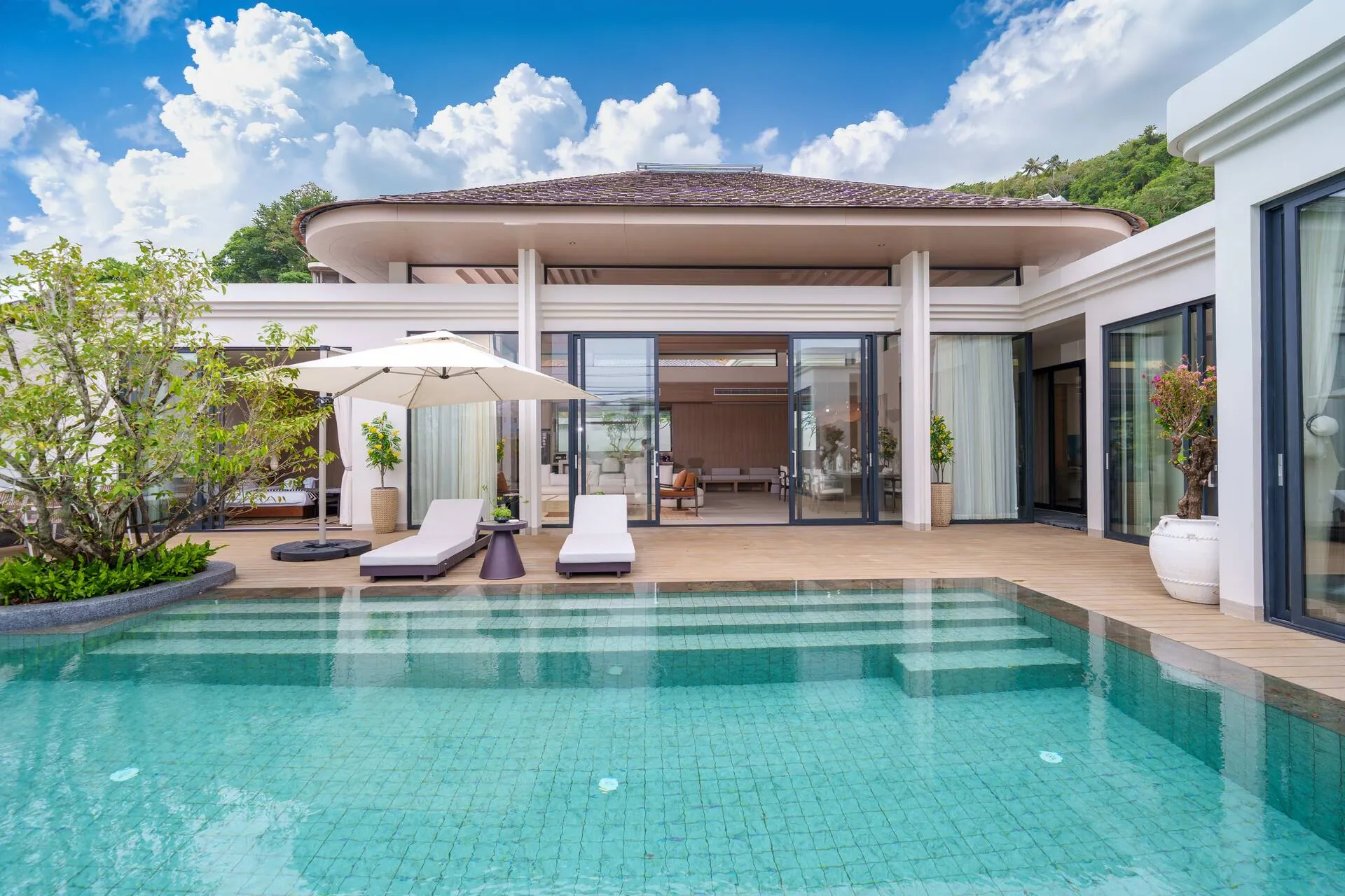 Private pool with sun loungers under a parasol in front of the villa's glass-walled pavilion, with a wooded hill behind