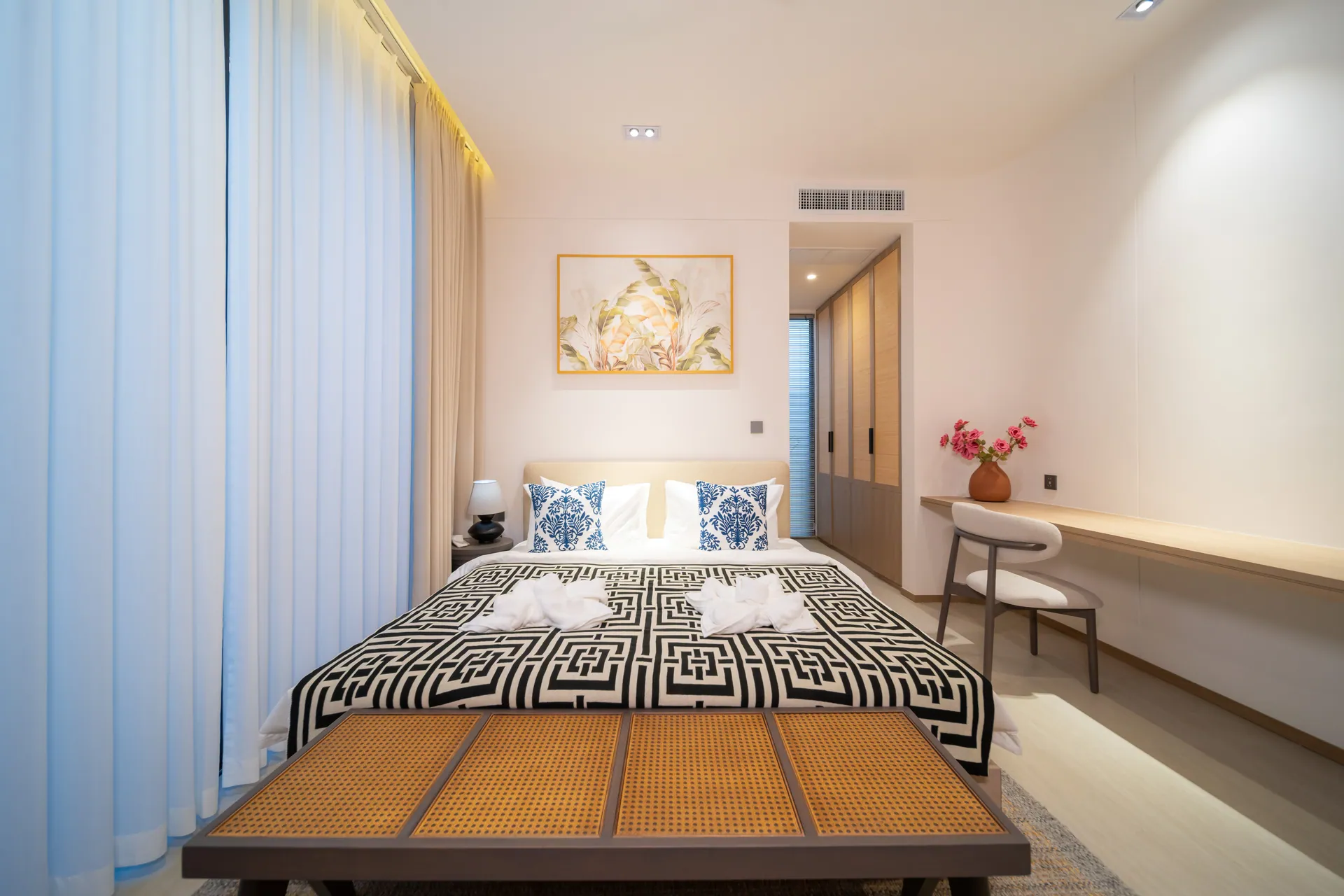 Bedroom with a king bed, blue patterned cushions and a writing desk beside the curtained window