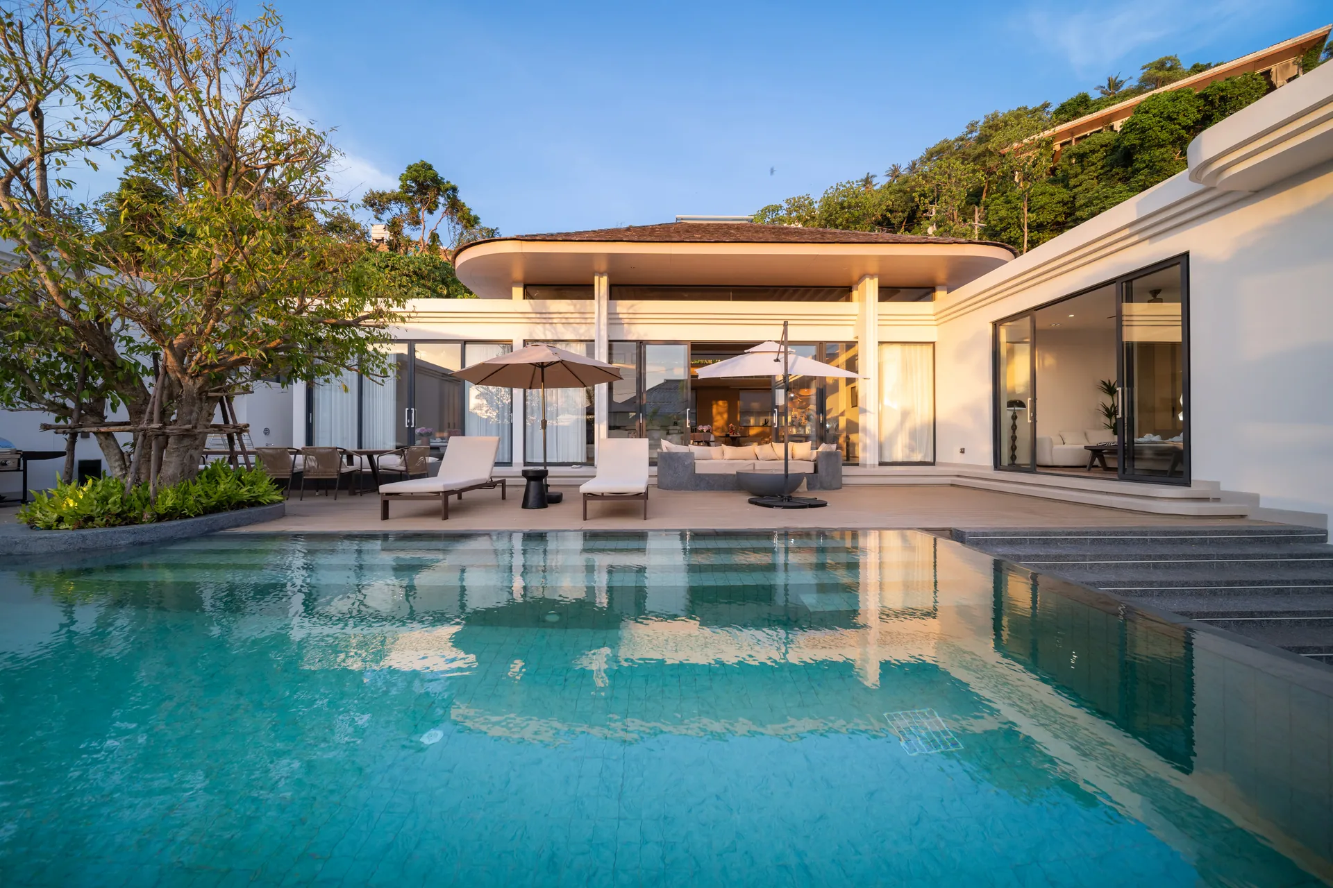Private pool and timber deck in late-afternoon sun, with parasols in front of the living pavilion and a wooded hillside behind