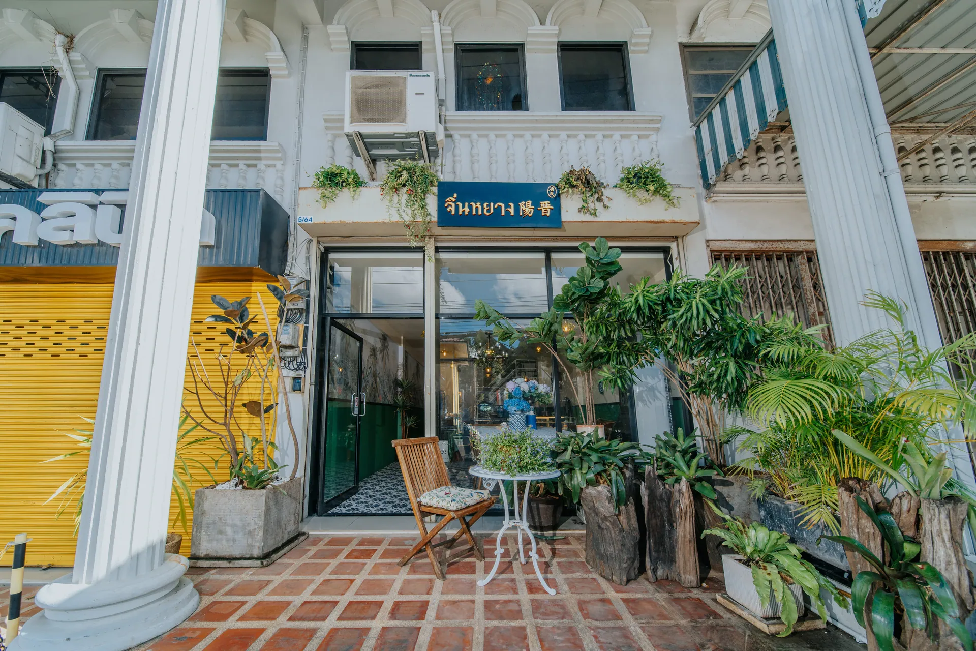 Tiled entrance terrace with a bistro table, a folding chair and tropical plants between the colonnade columns
