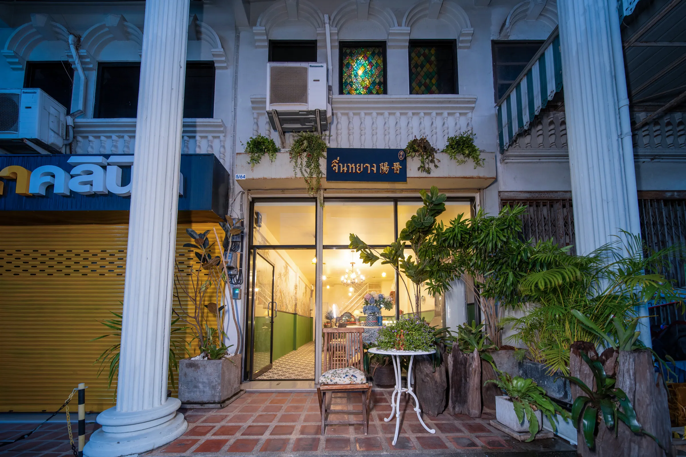 Shophouse front at dusk, with white columns, potted palms and the lit glass entrance to the ground-floor café