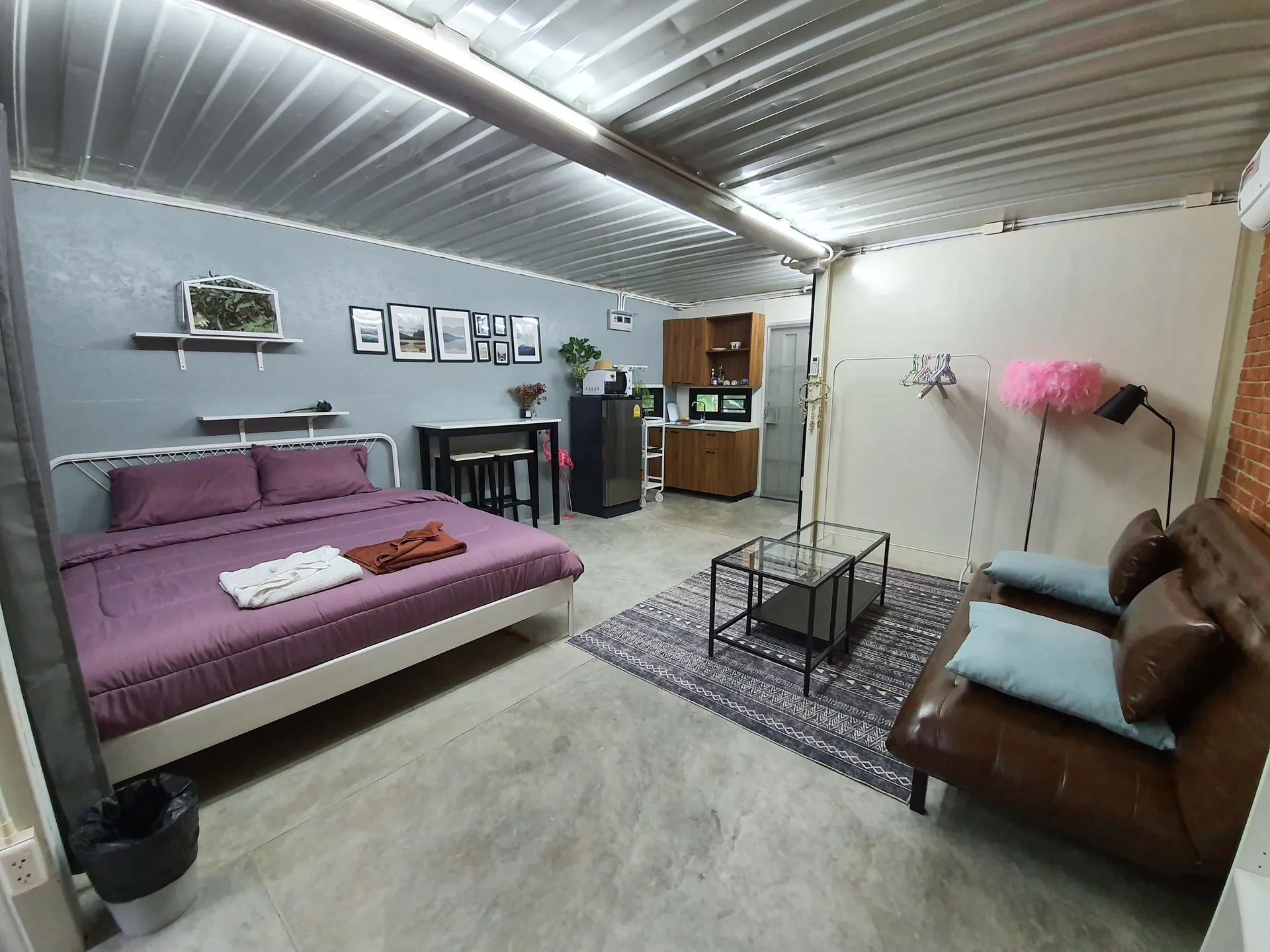 Studio bedroom with plum linen, a lounge corner and a kitchenette under a corrugated steel ceiling