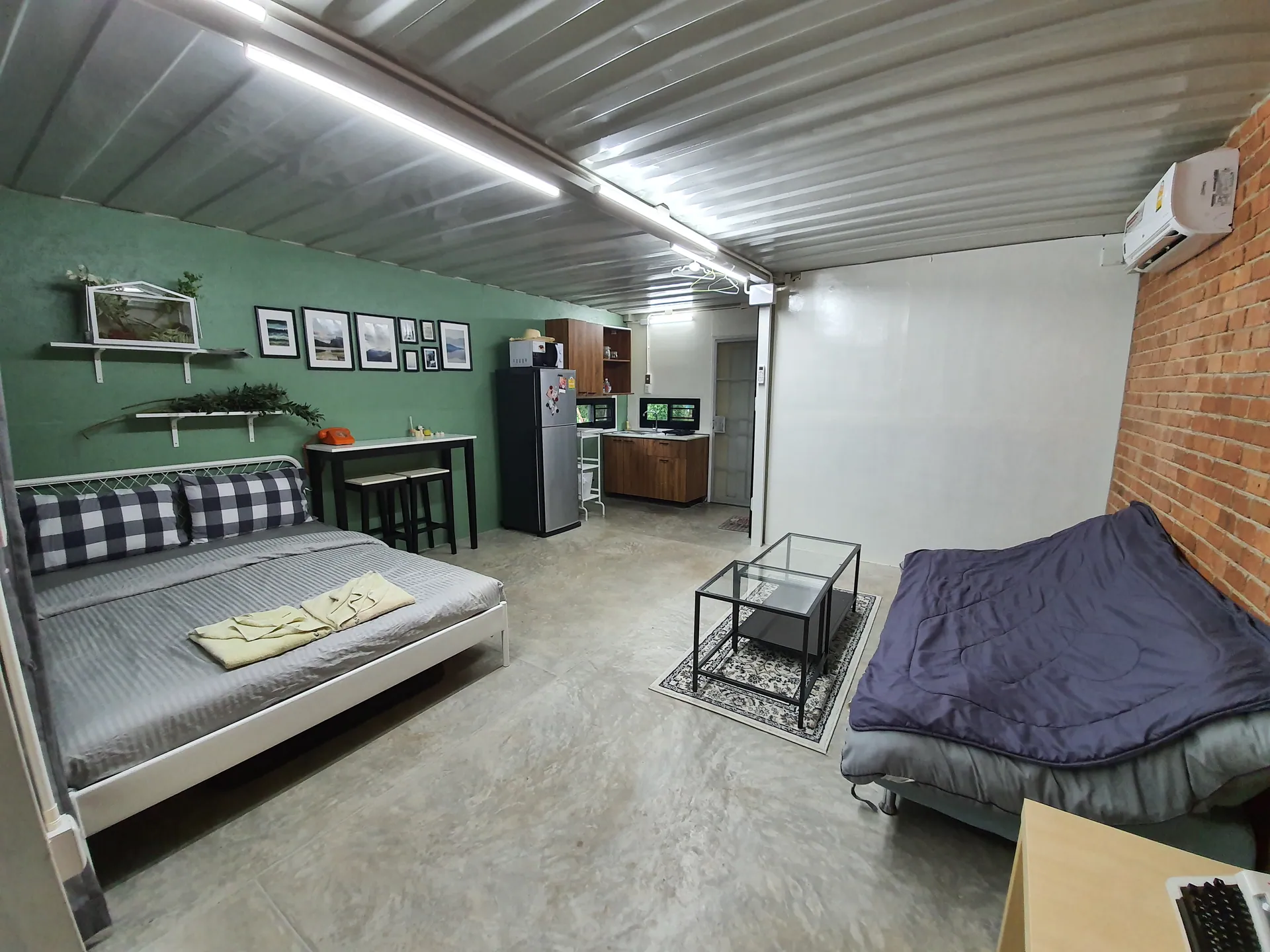 Bedroom with checked pillows, a green wall of framed prints and a kitchenette at the far end