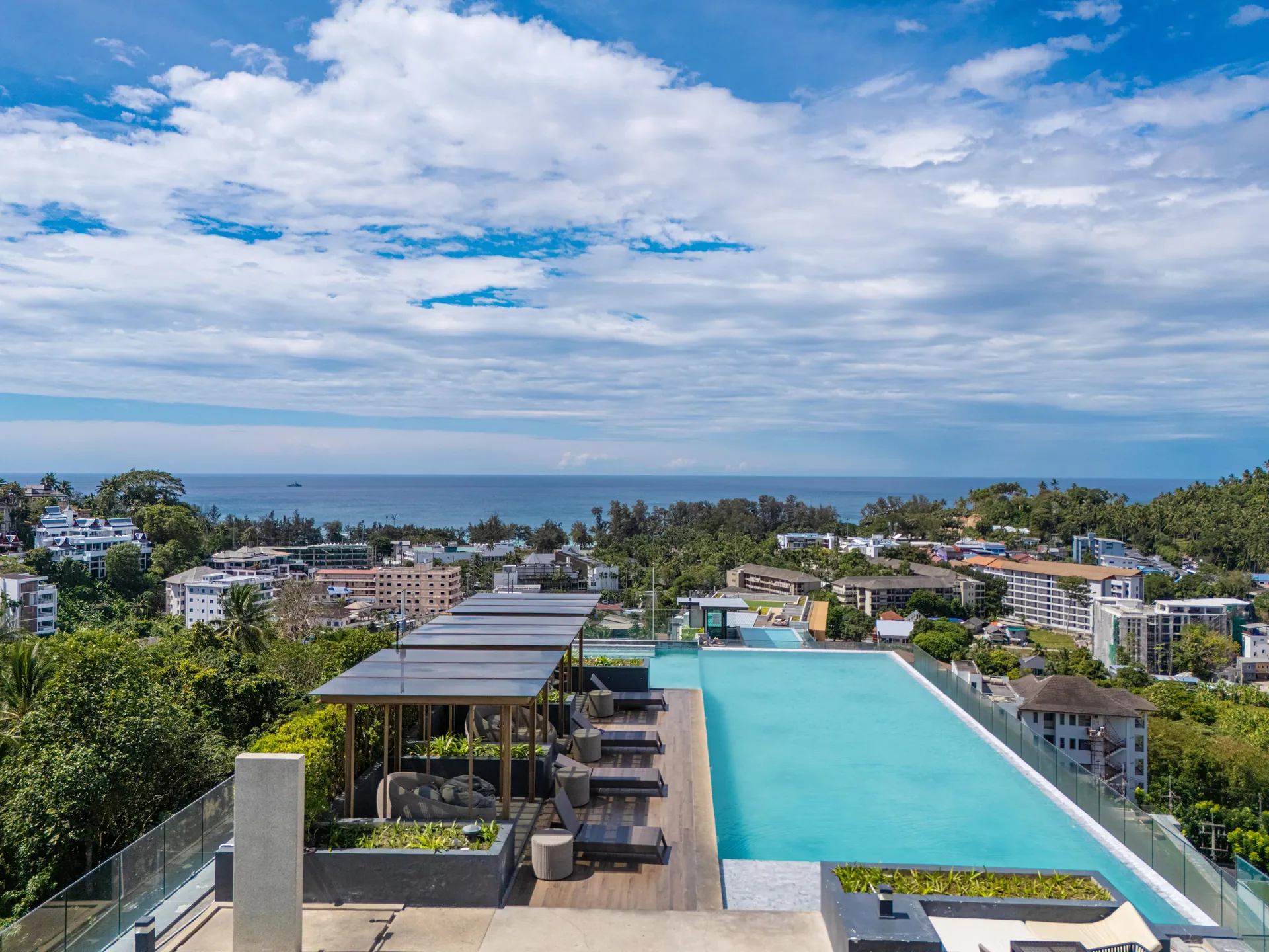 Rooftop infinity pool with shaded daybeds and sun loungers, looking out over the treetops to the Andaman Sea