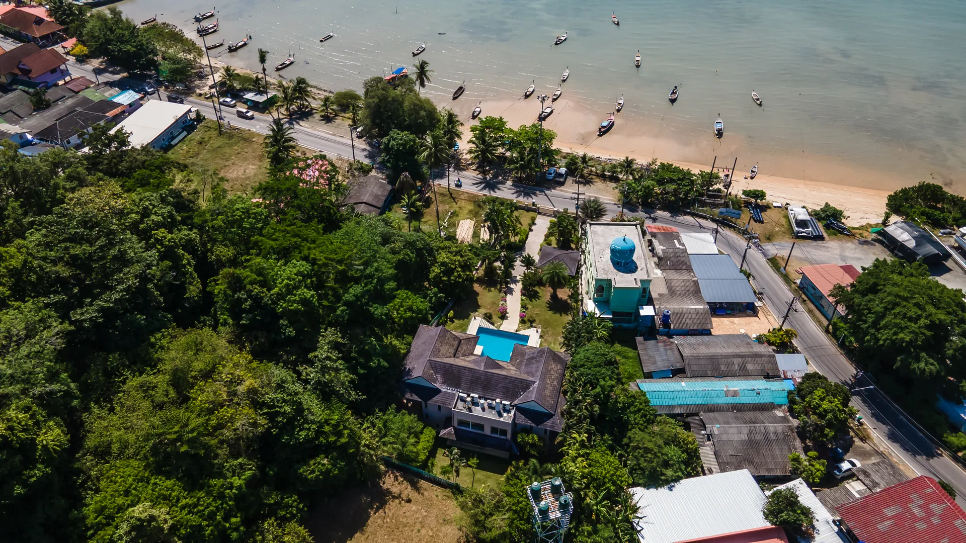 Aerial view of the villa in its wooded plot, with the coast road and a beach lined with longtail boats beyond