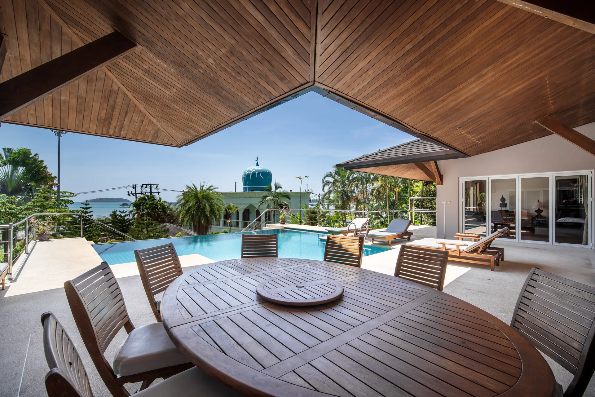 Oval teak dining table under the deep eaves beside the pool, with the bay visible beyond the palms