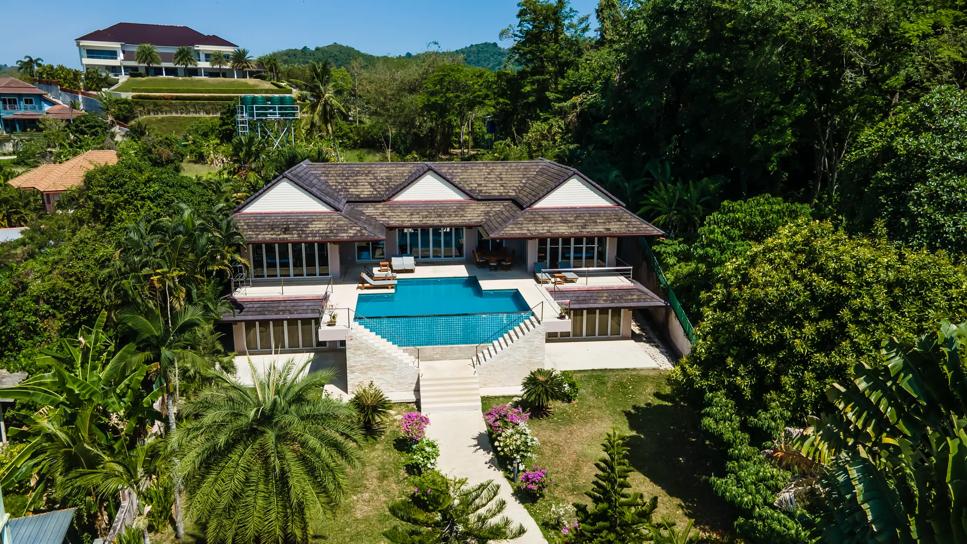 Aerial view of the villa among palms and tall trees, with a planted garden and steps rising to the pool