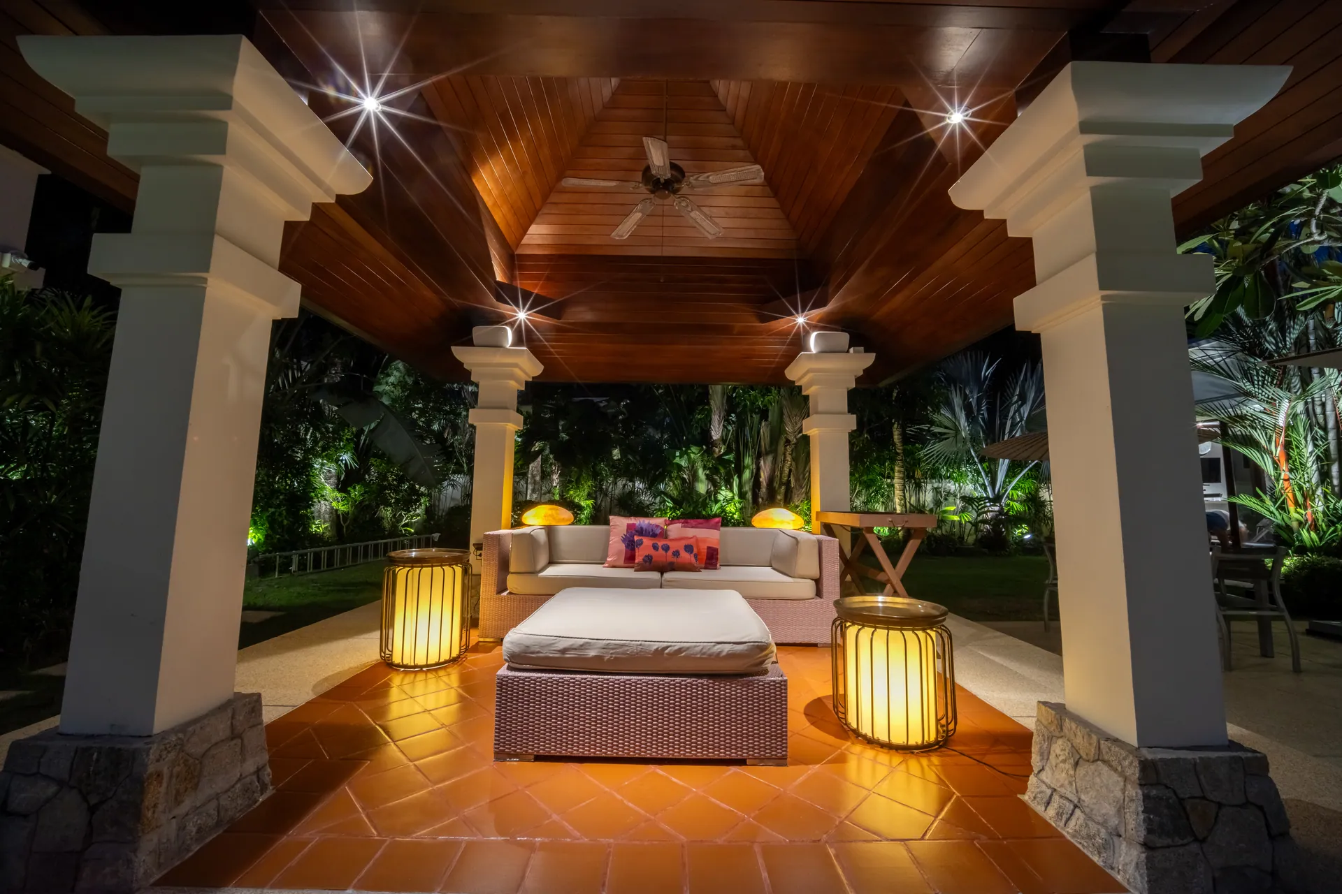 Columned garden sala with a cushioned daybed and floor lanterns, lit in the evening