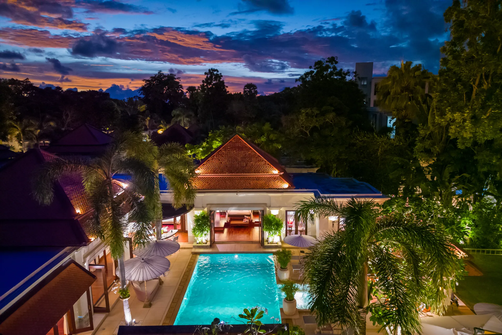Aerial view of the villa at dusk, its lit pool framed by pavilion roofs and tropical trees