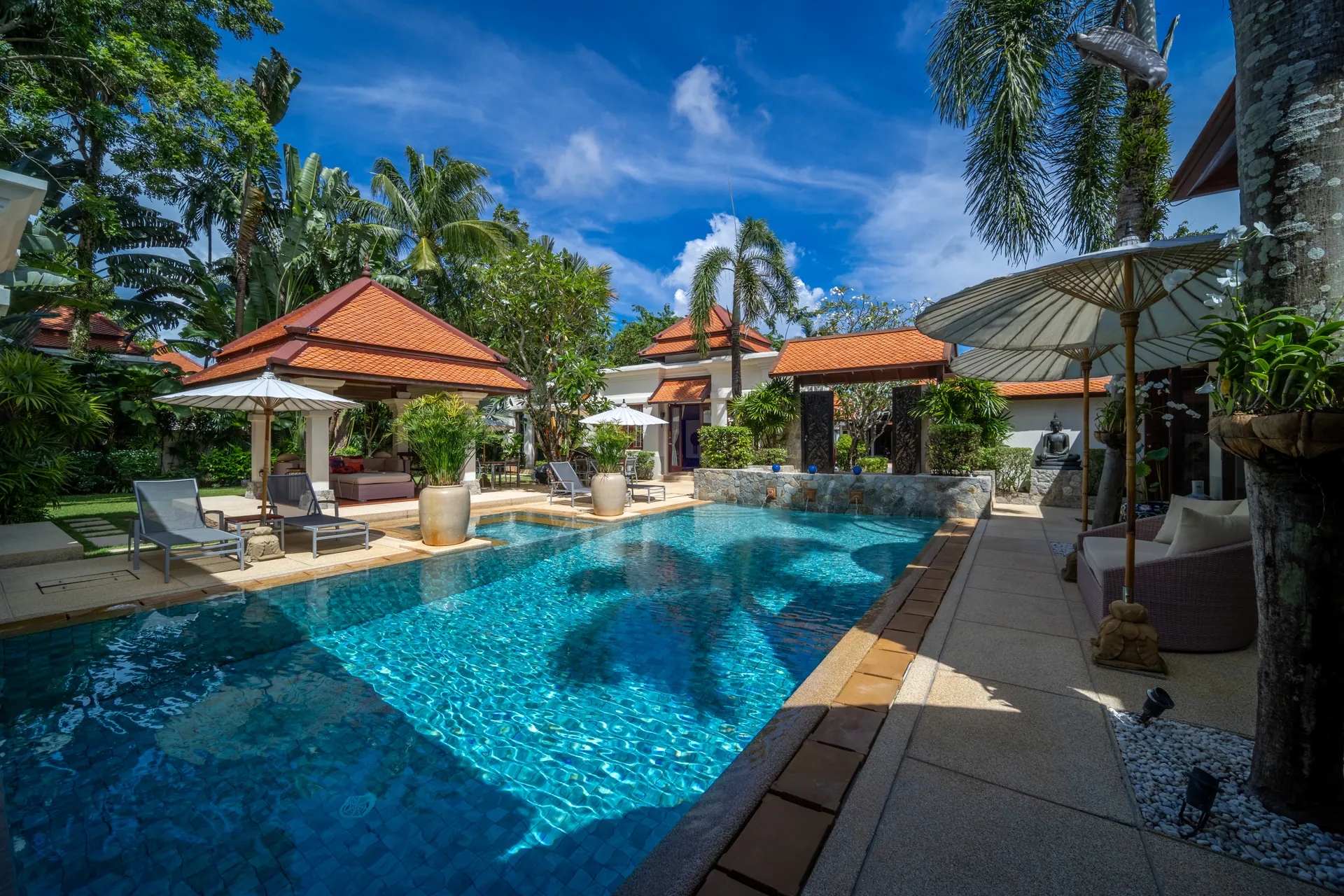 Long tiled pool in daylight, lined with loungers, parasols and terracotta-roofed pavilions