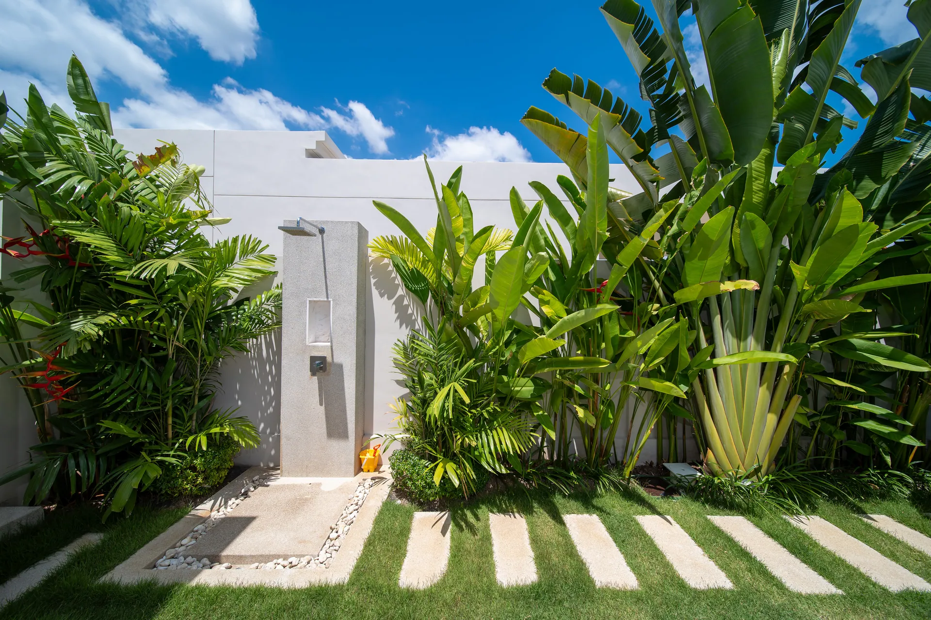 Outdoor shower against a white garden wall, reached by stepping stones across the lawn among banana plants