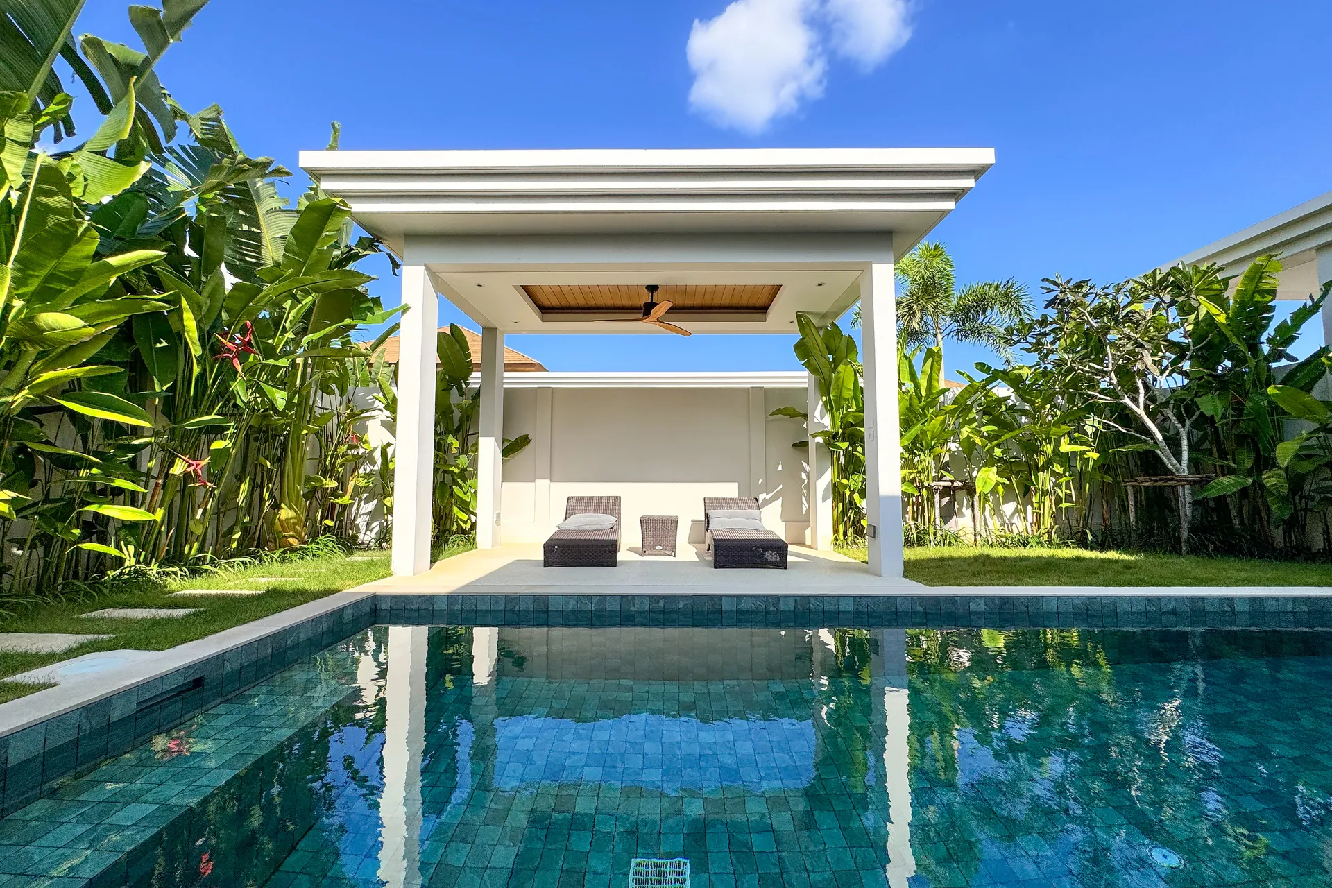 Covered pool sala with two loungers, framed by banana plants across the water