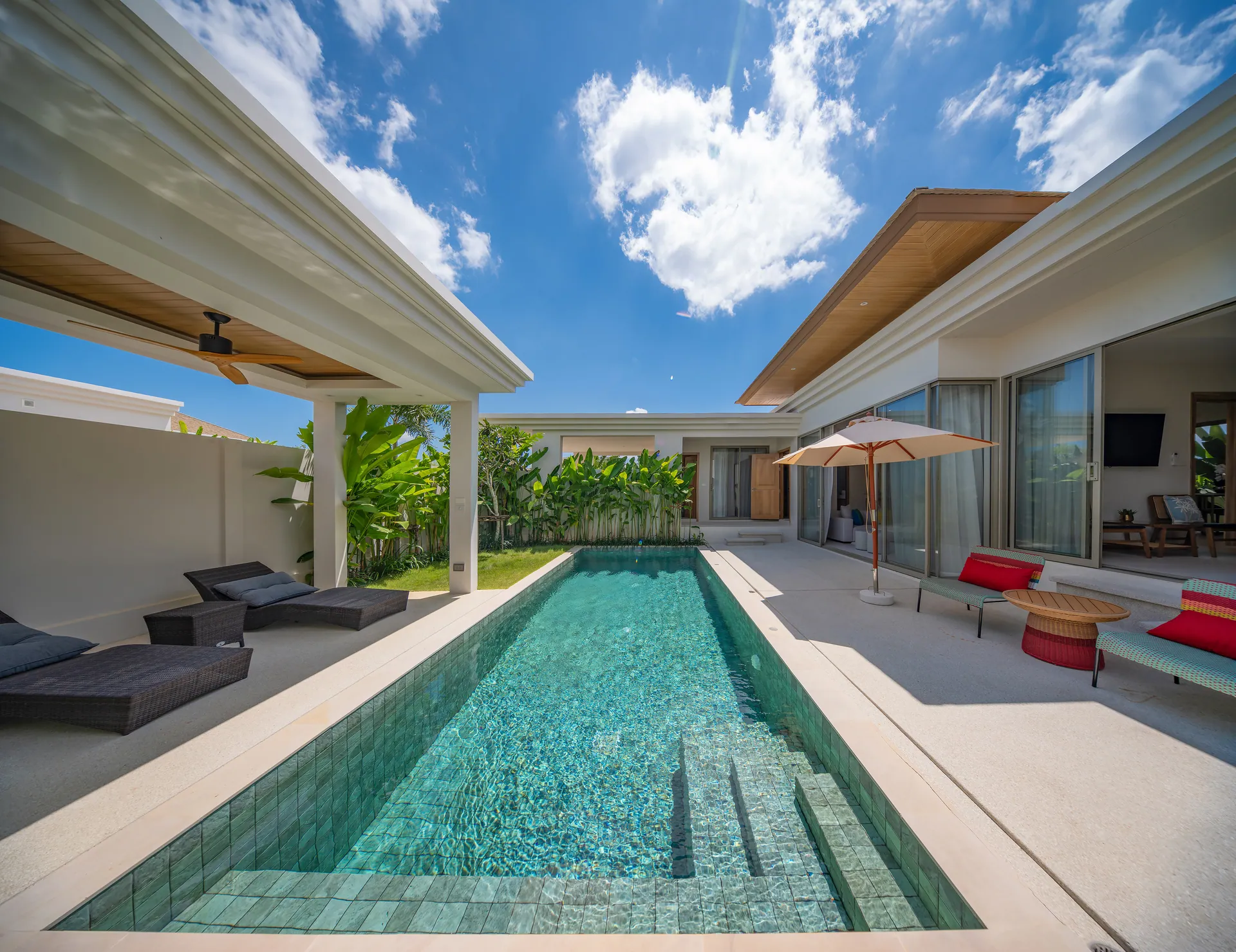 Long tiled pool beside the terrace, with loungers on one side and the villa's glass doors and red-cushioned chairs on the other