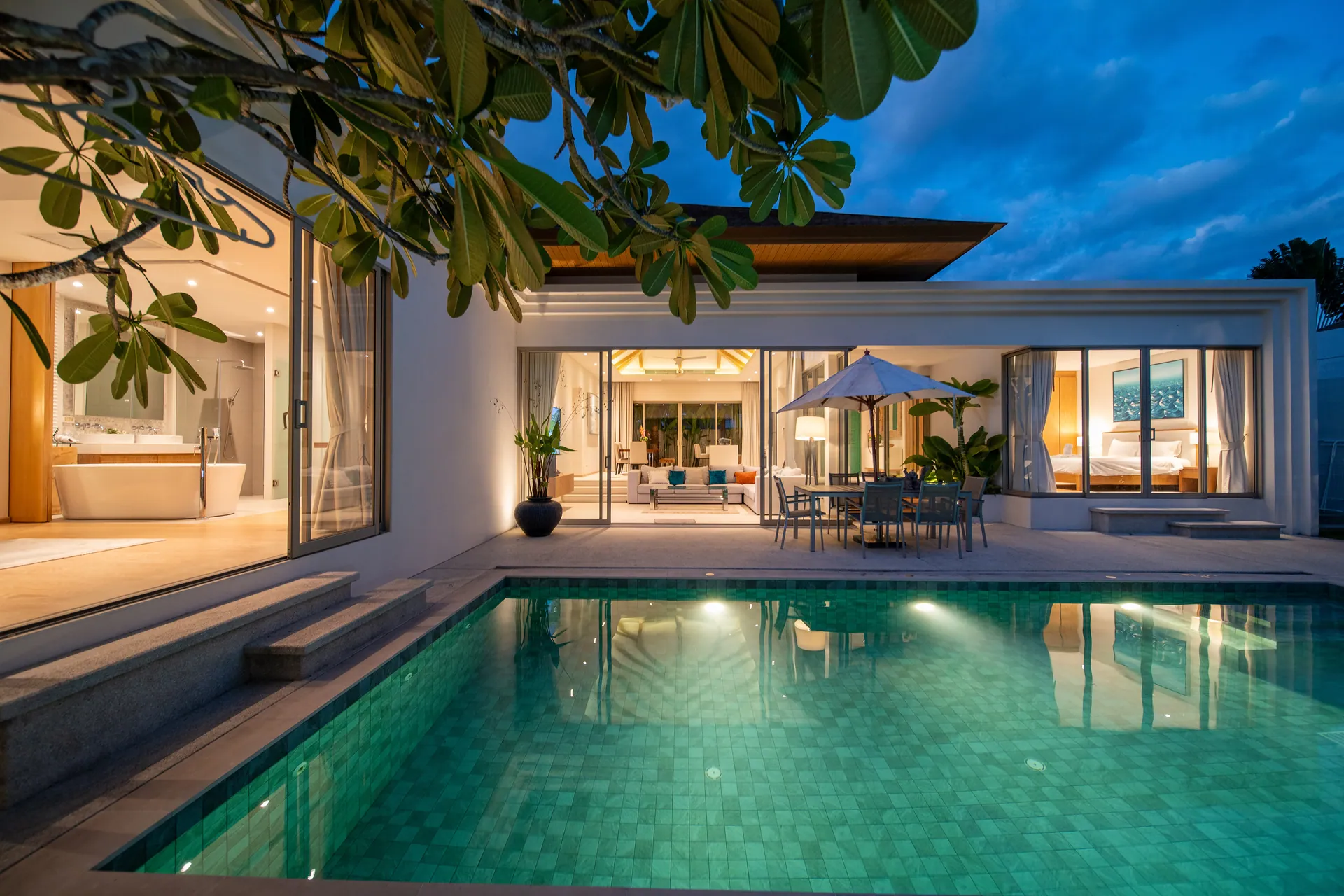 Private pool at dusk, lit from below, with the single-storey villa and its open living room beyond
