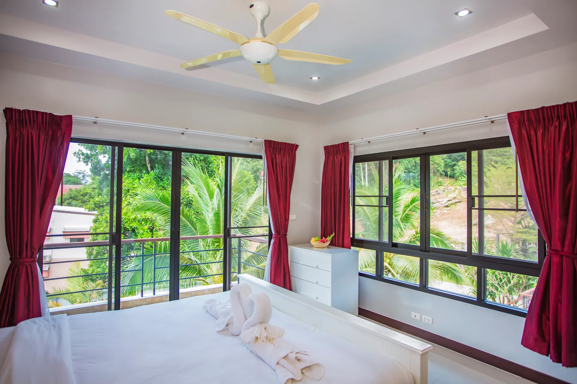 Corner bedroom with red curtains, a ceiling fan and glass doors to a balcony over the palms