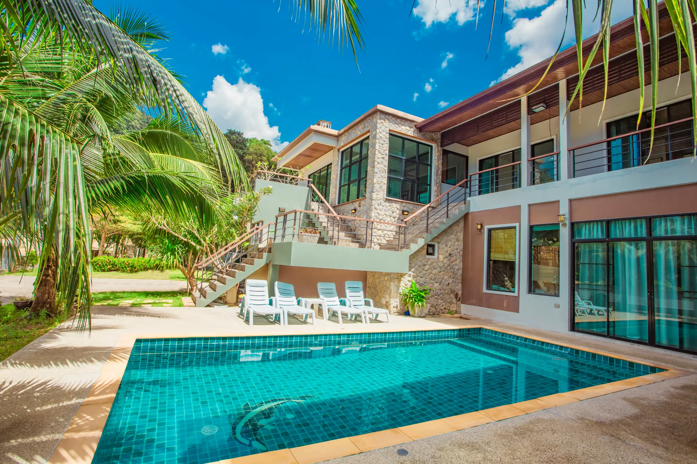 Private pool in front of the two-storey villa, with a stone-clad wing, exterior stairs, sun loungers and coconut palms