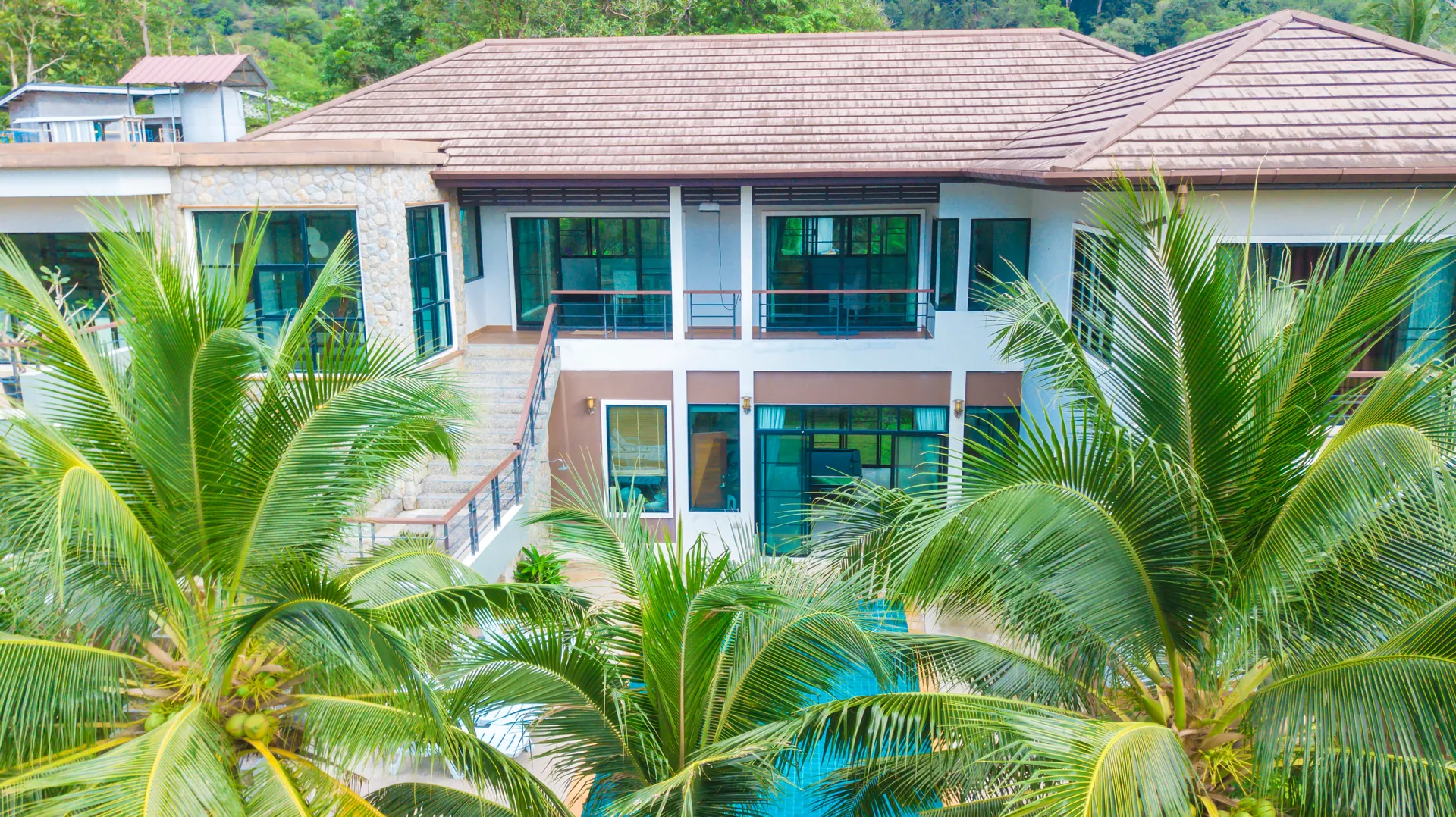 Aerial view of the villa's tiled roofs and stone-clad facade above a canopy of coconut palms