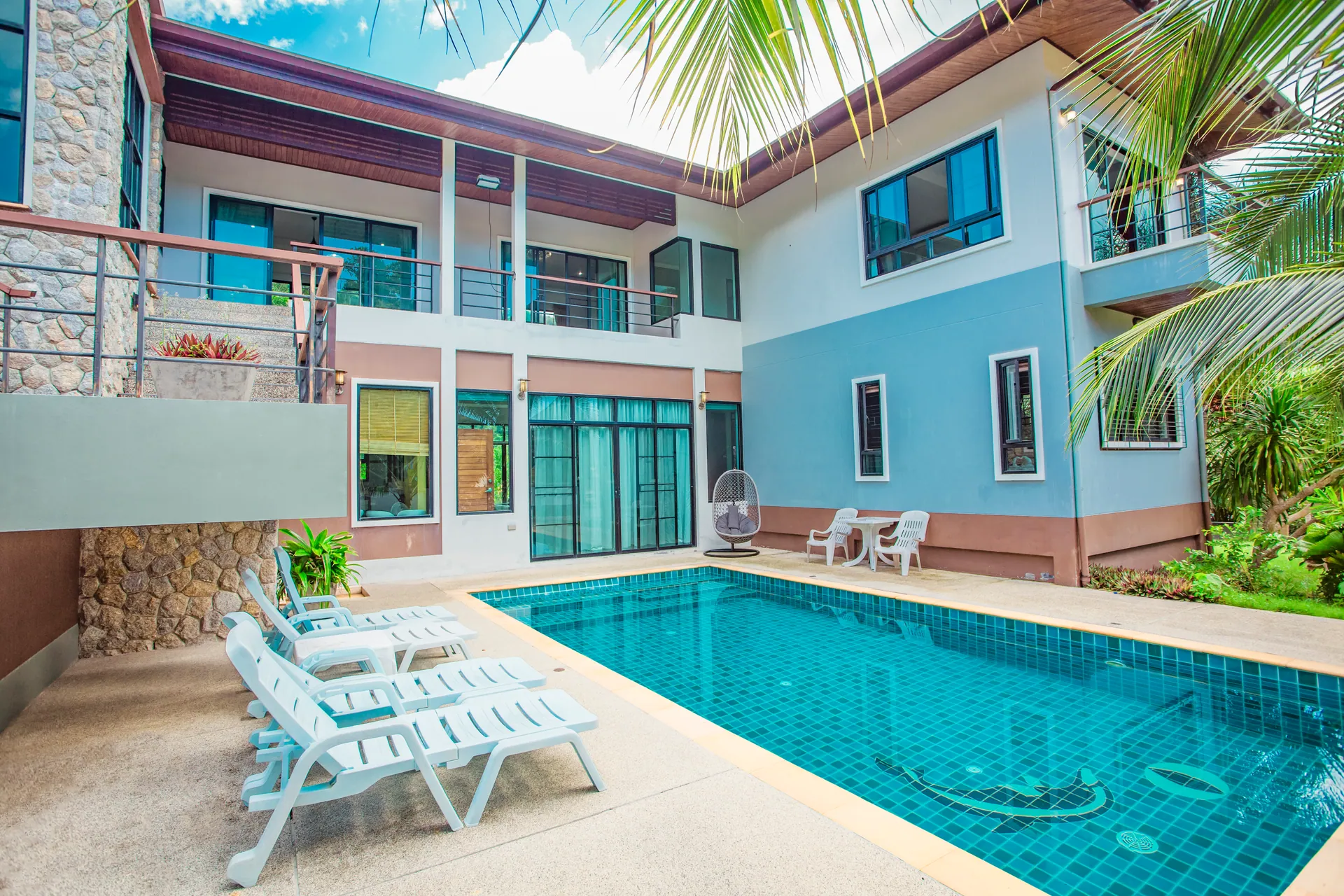 L-shaped villa wrapped around the pool, with white sun loungers on the deck and a hanging egg chair by the glass doors