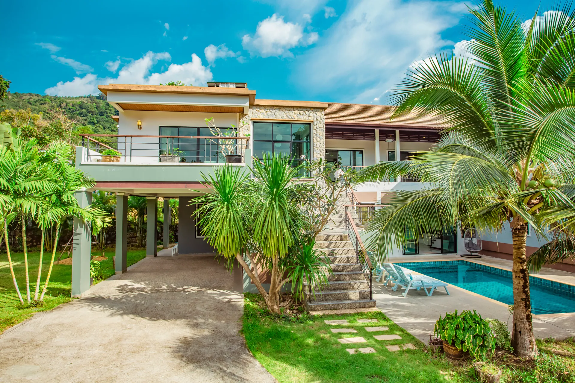 Front of the villa with a driveway, a lawn with stepping stones, palms and a glimpse of the pool