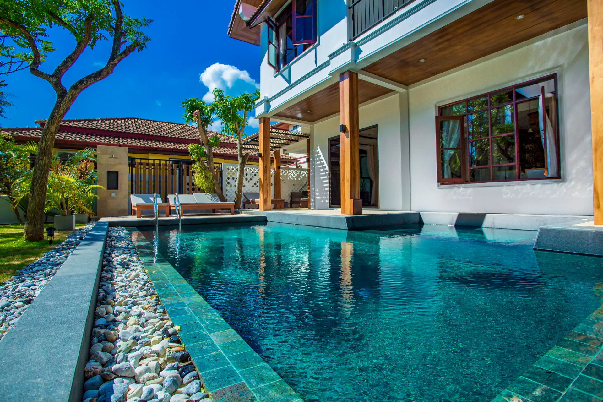 Private pool running alongside the villa, edged with pebbles and green stone tiles, with sun loungers beyond