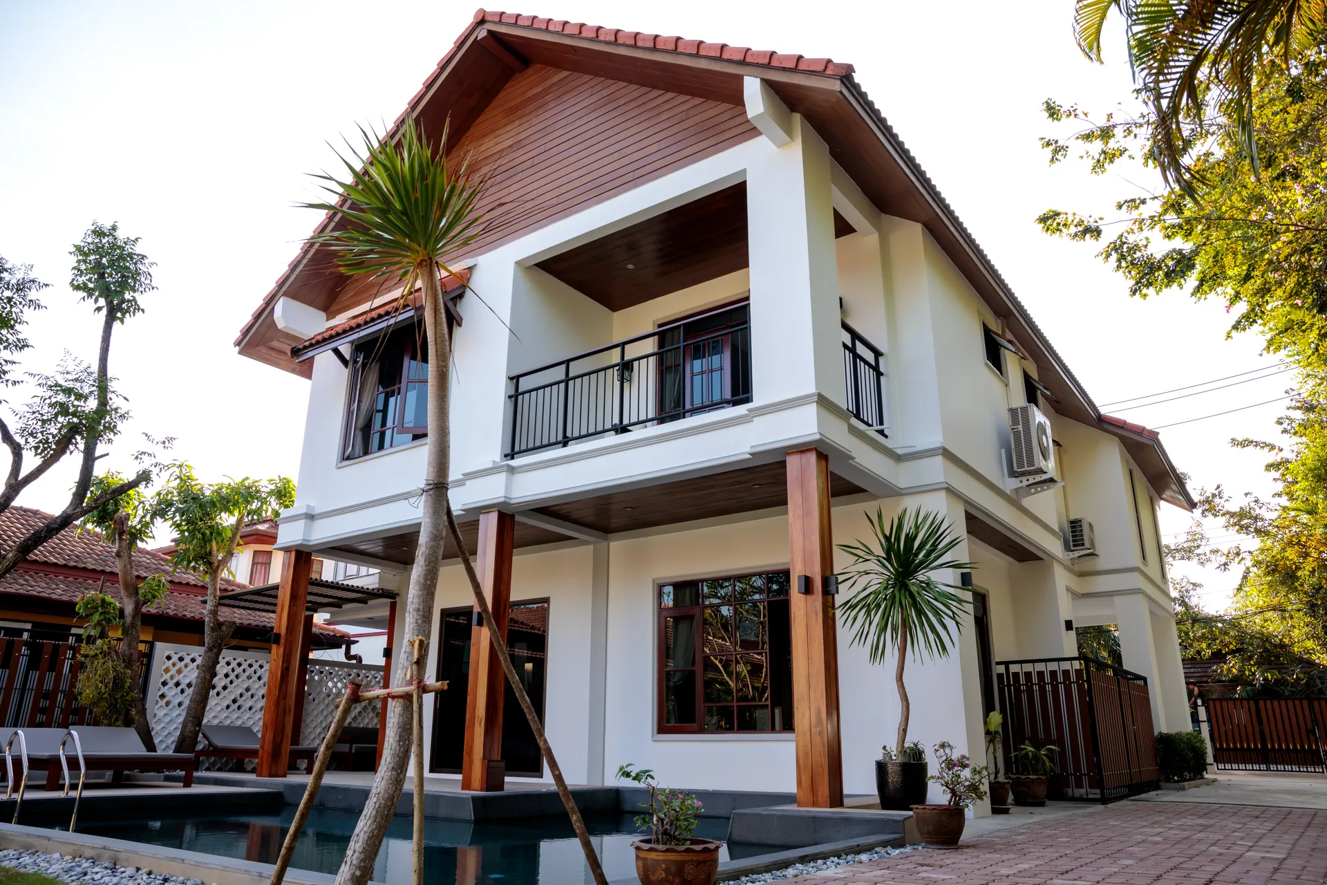 Villa facade with an upstairs balcony, timber columns and potted palms at the edge of the pool