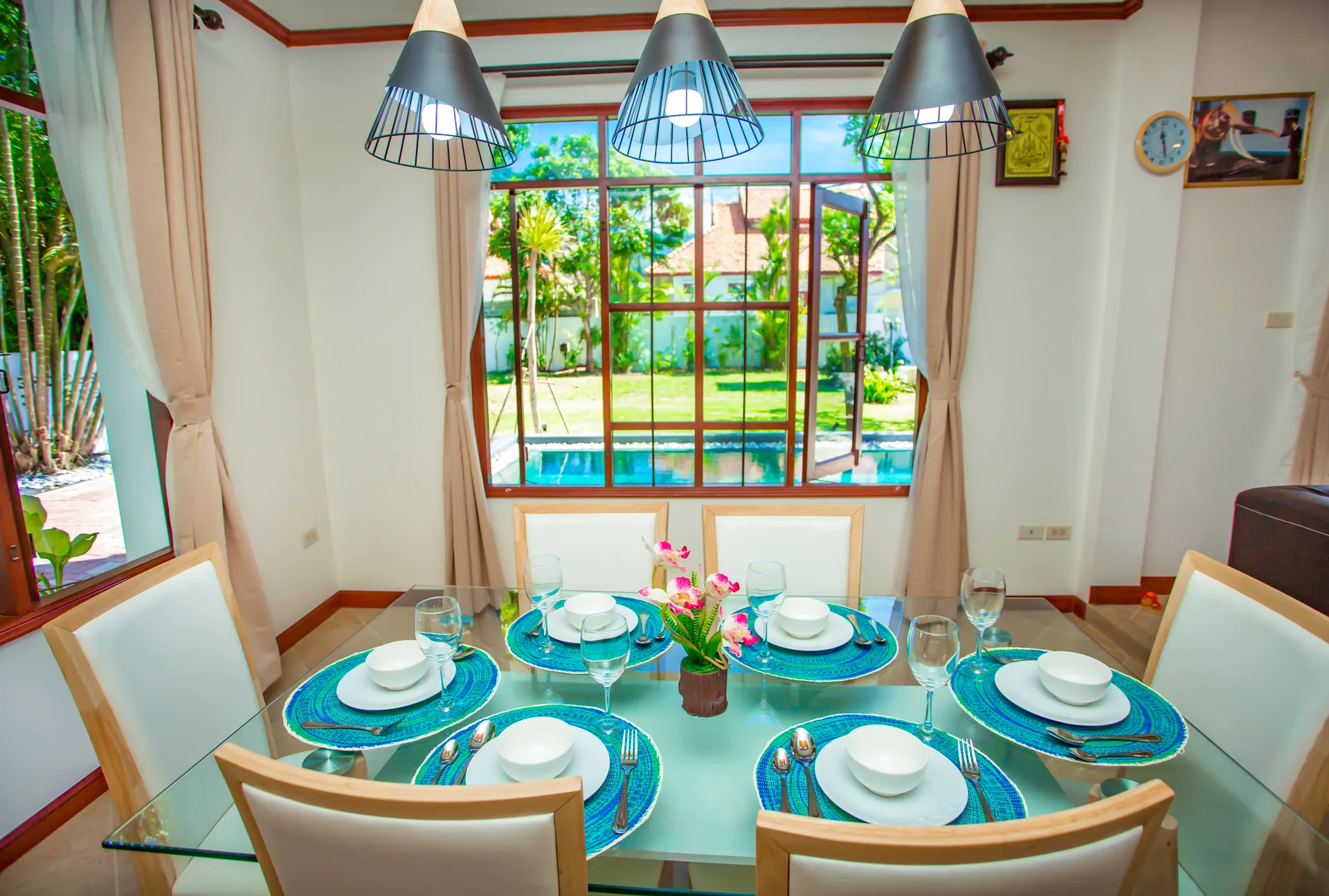 Glass dining table set for six below three pendant lights, with a window onto the pool and garden