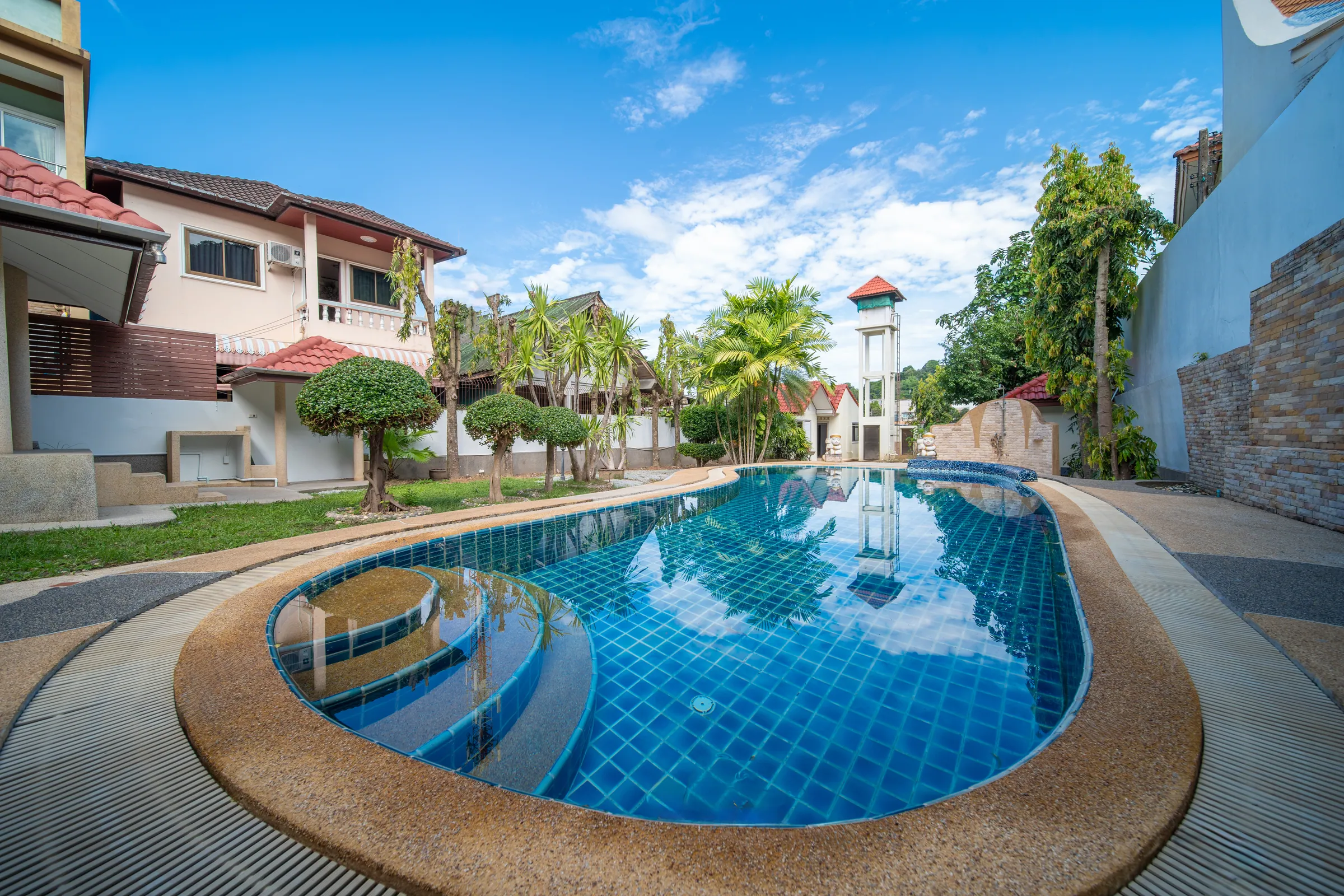 Long freeform pool with curved entry steps, framed by a lawn, clipped trees and palms under a blue sky