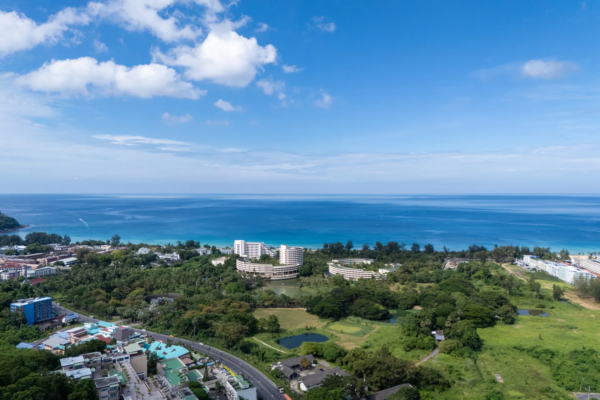 Aerial view over Karon, with green open land and beachfront hotels in front of the Andaman Sea