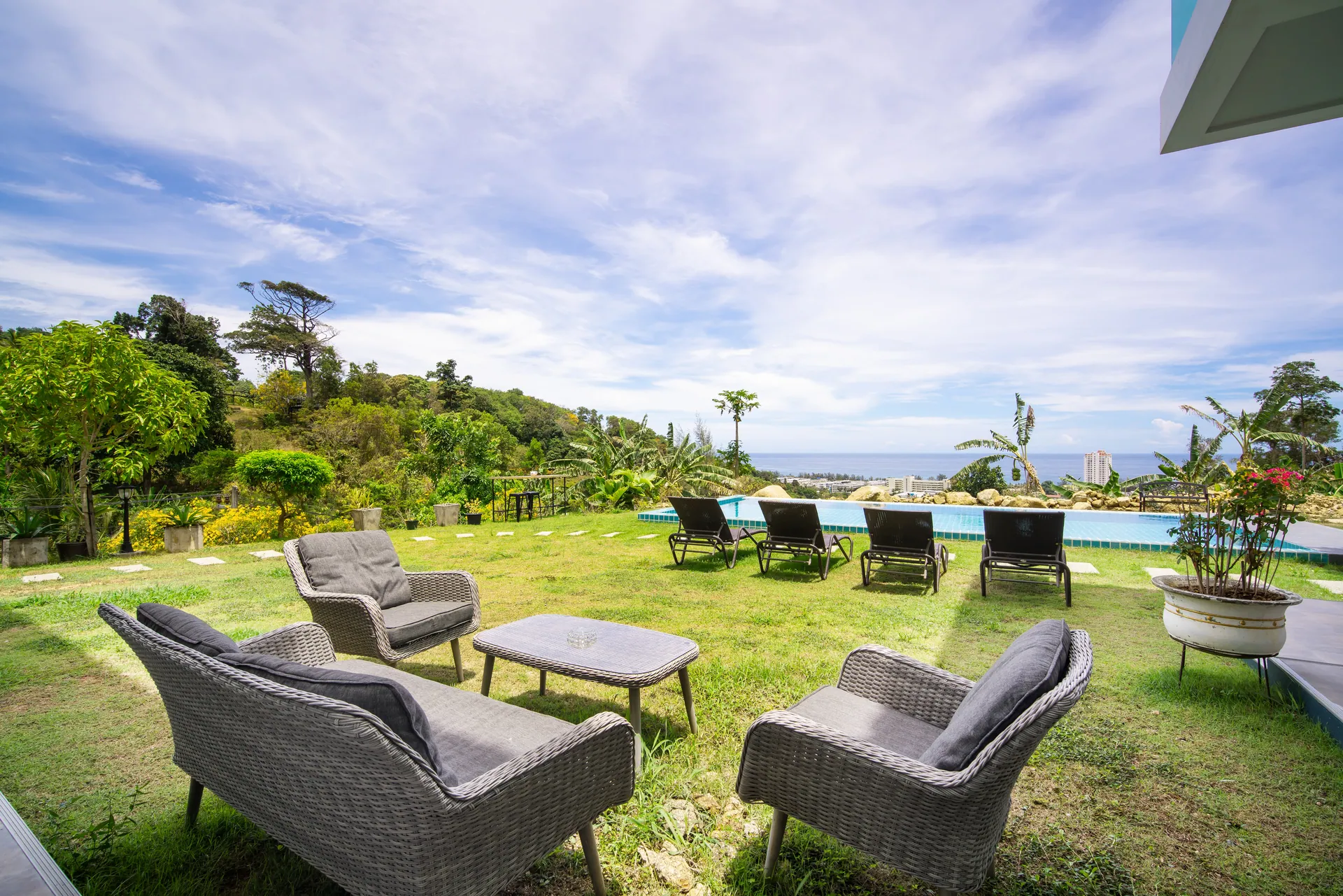 Wicker lounge chairs on the lawn, with the pool, a row of sun loungers and a distant sea view beyond