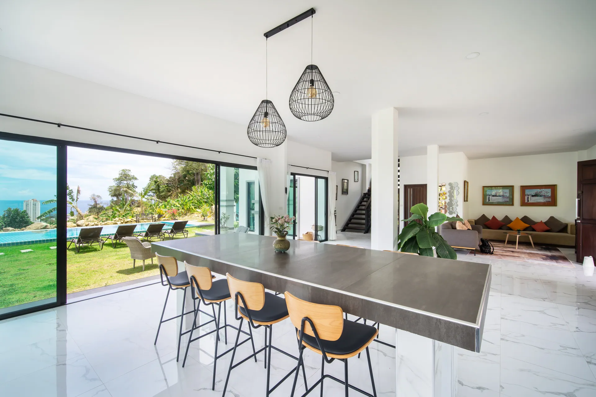Open-plan dining area with a long bar-height table and high stools, beside glass doors to the pool lawn