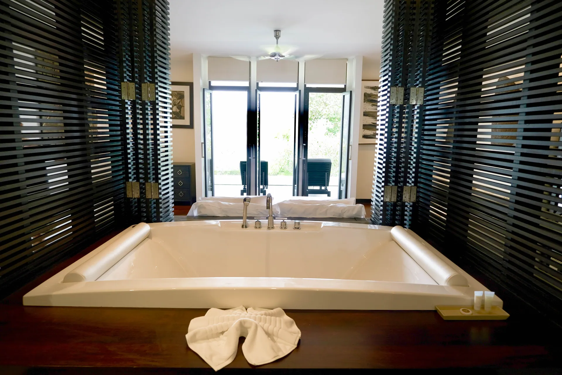 Deep soaking tub set between slatted timber screens, with the bedroom beyond