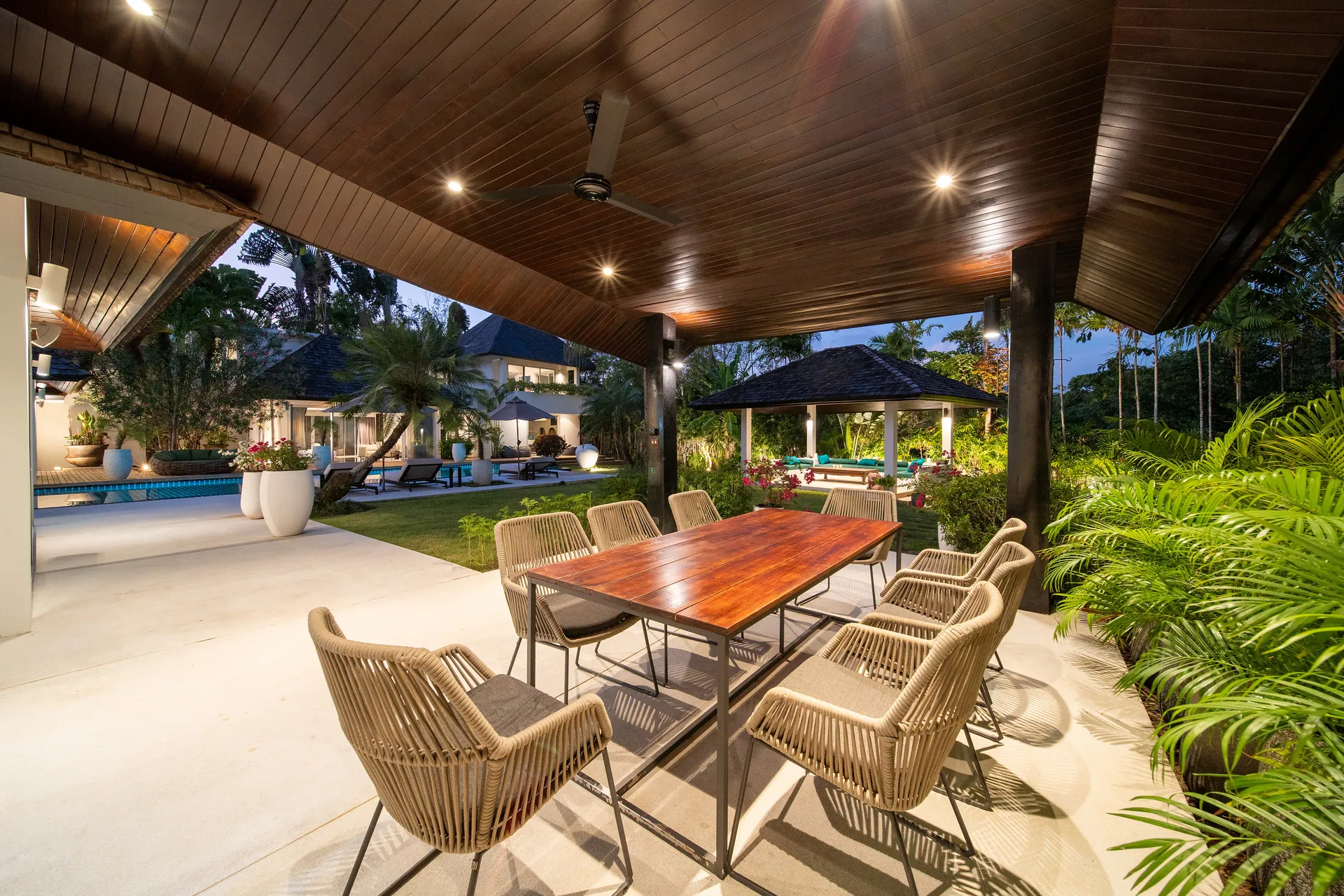 Covered outdoor dining terrace with a timber table and woven chairs, lit in the evening