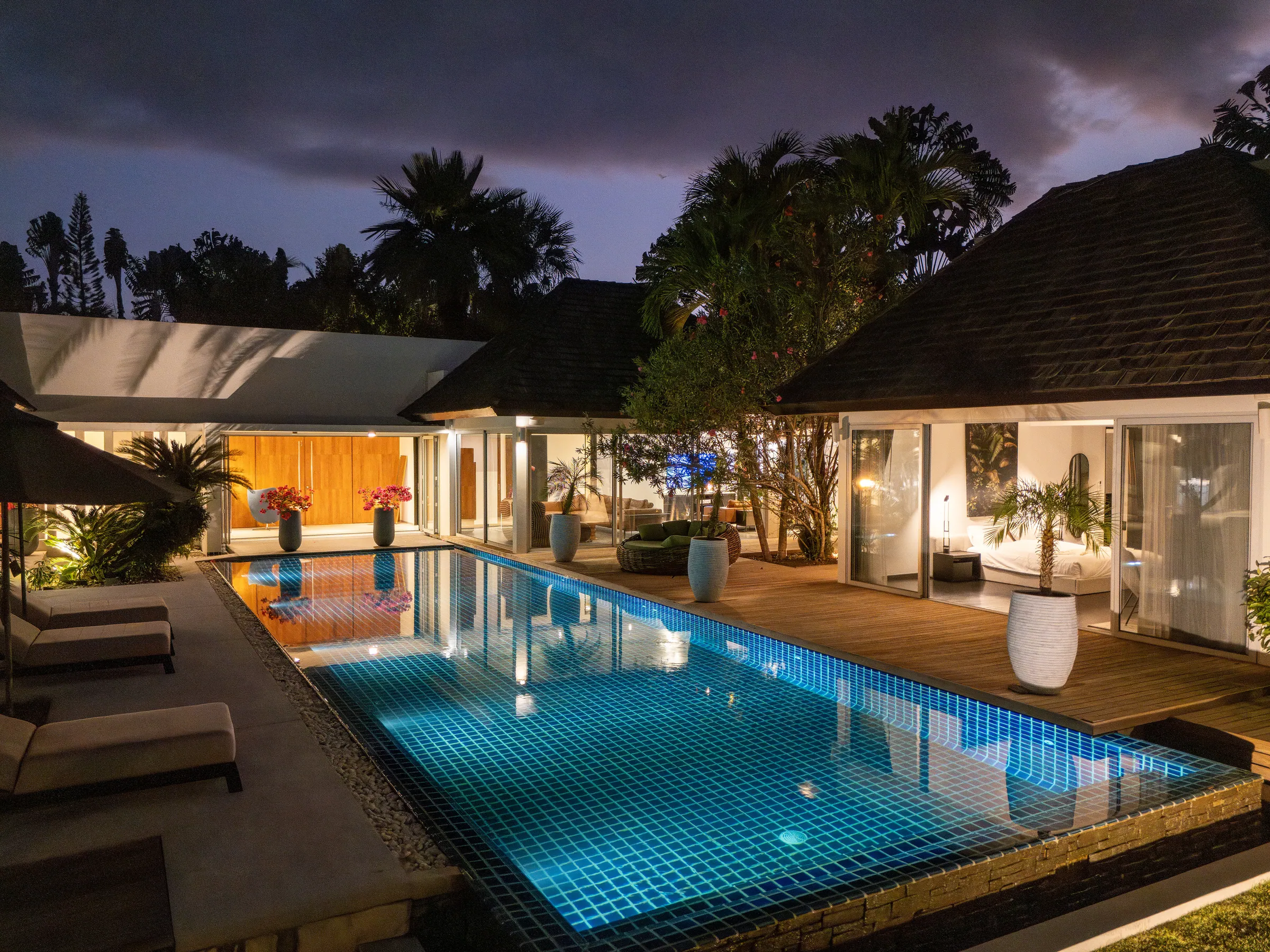 Long tiled pool lit at dusk, with a timber deck, sun loungers and glass-fronted pavilions glowing behind