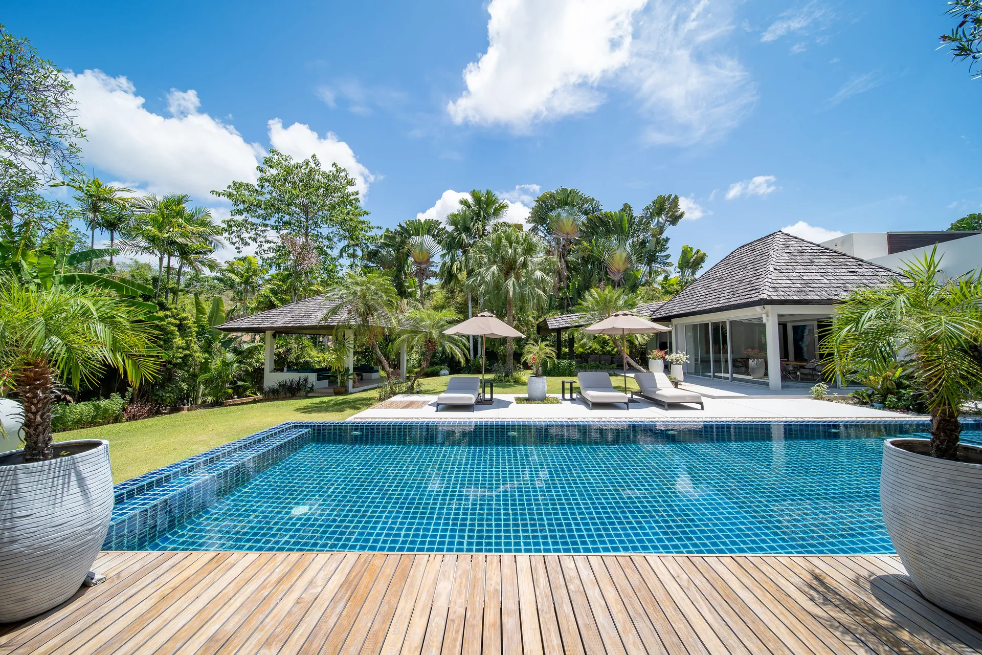 The pool seen from the timber deck, with loungers under parasols and a pavilion across the lawn