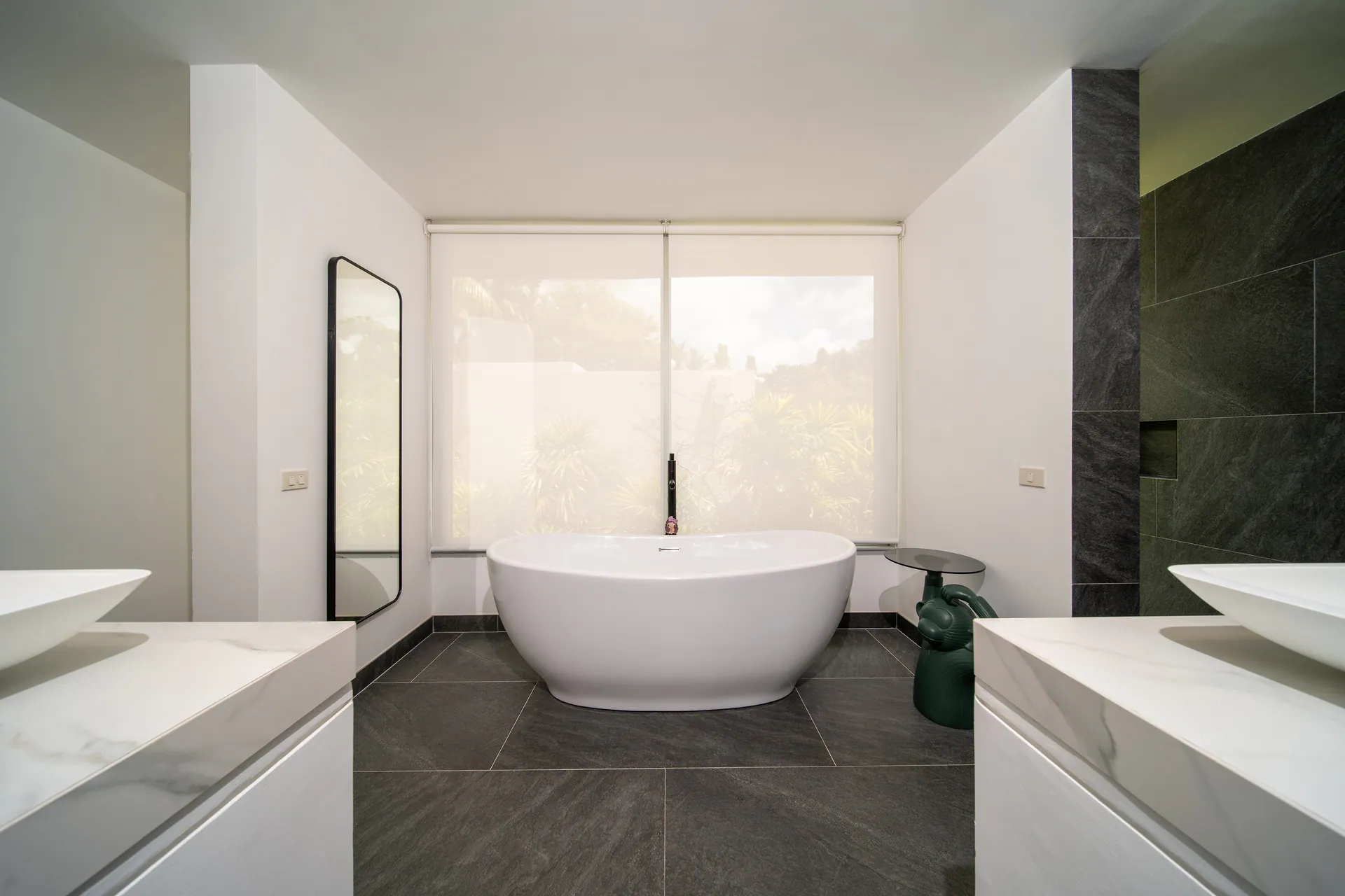 Bathroom with a freestanding bath beneath a screened window, between two marble-topped vanities