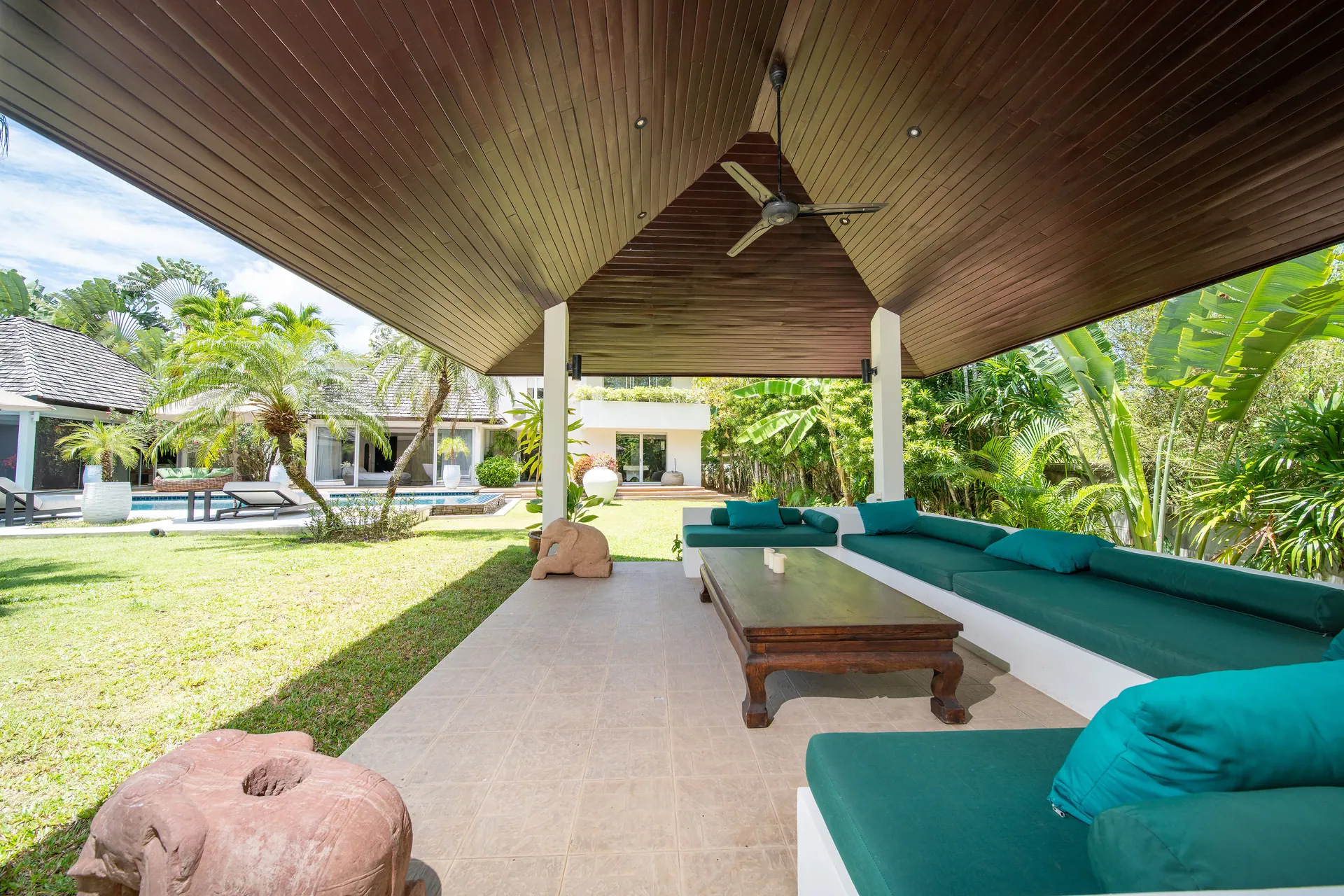 Open-sided garden sala with built-in cushioned seating and a low timber table, looking across the lawn