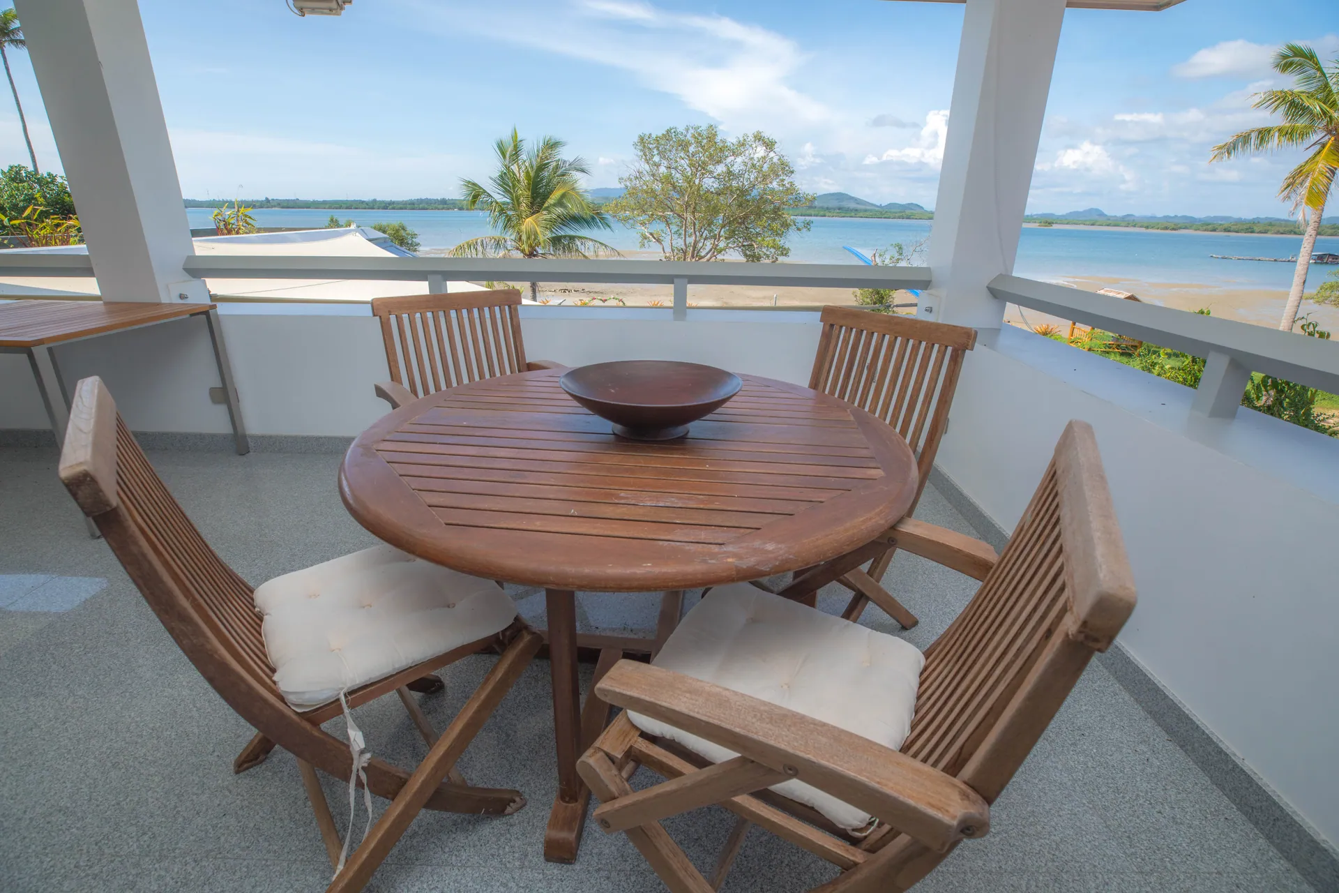 Balcony with a round teak table and folding chairs, overlooking the garden and the bay