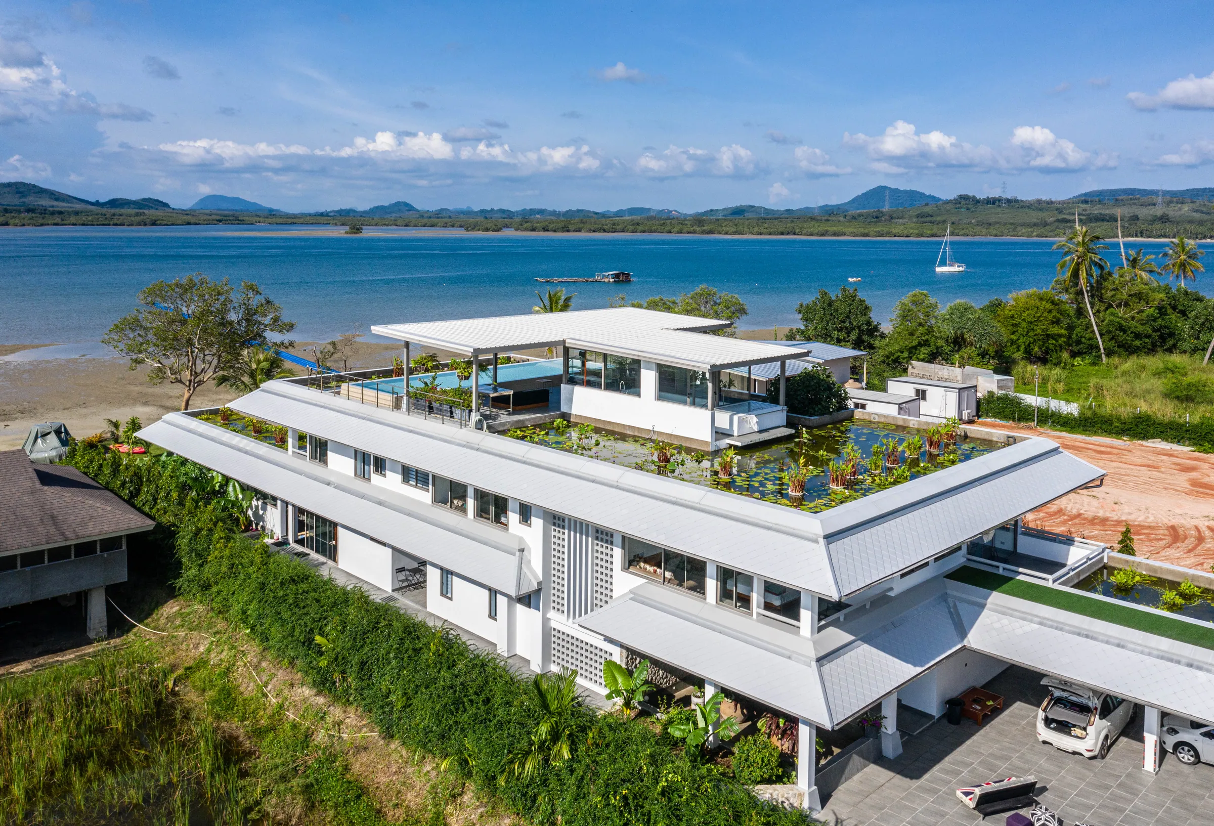 Aerial view of the white, tiered villa with its rooftop pool and water-lily roof gardens, with a calm bay and wooded hills beyond