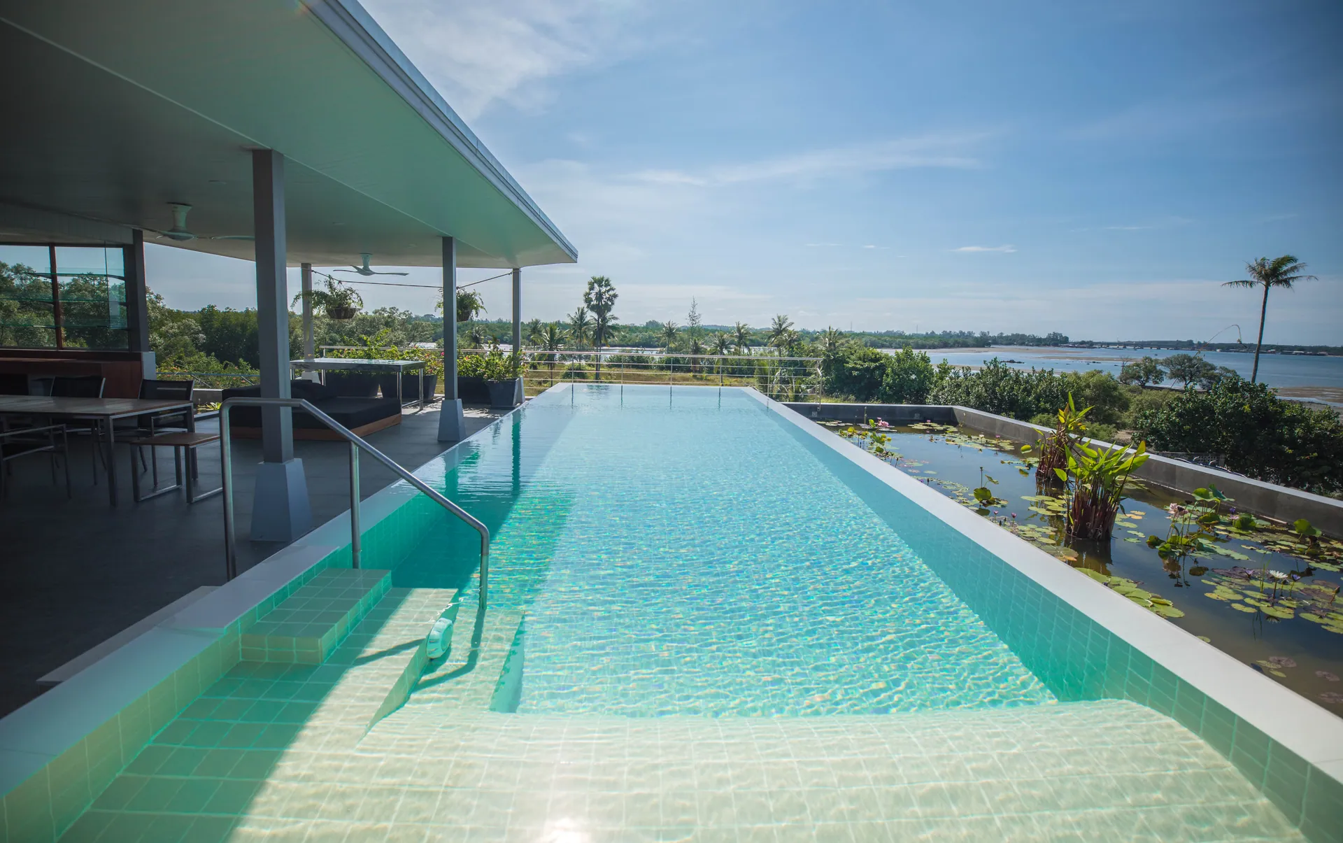 Rooftop infinity pool beside a covered pavilion, with a water-lily pond alongside and the bay in the distance