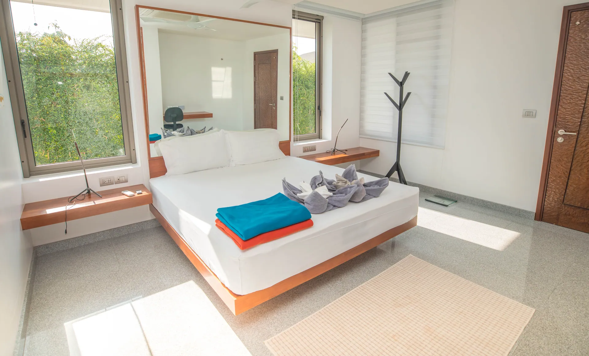 Bedroom with a platform bed, a teal throw, a wall mirror and a carved timber door, with windows onto greenery
